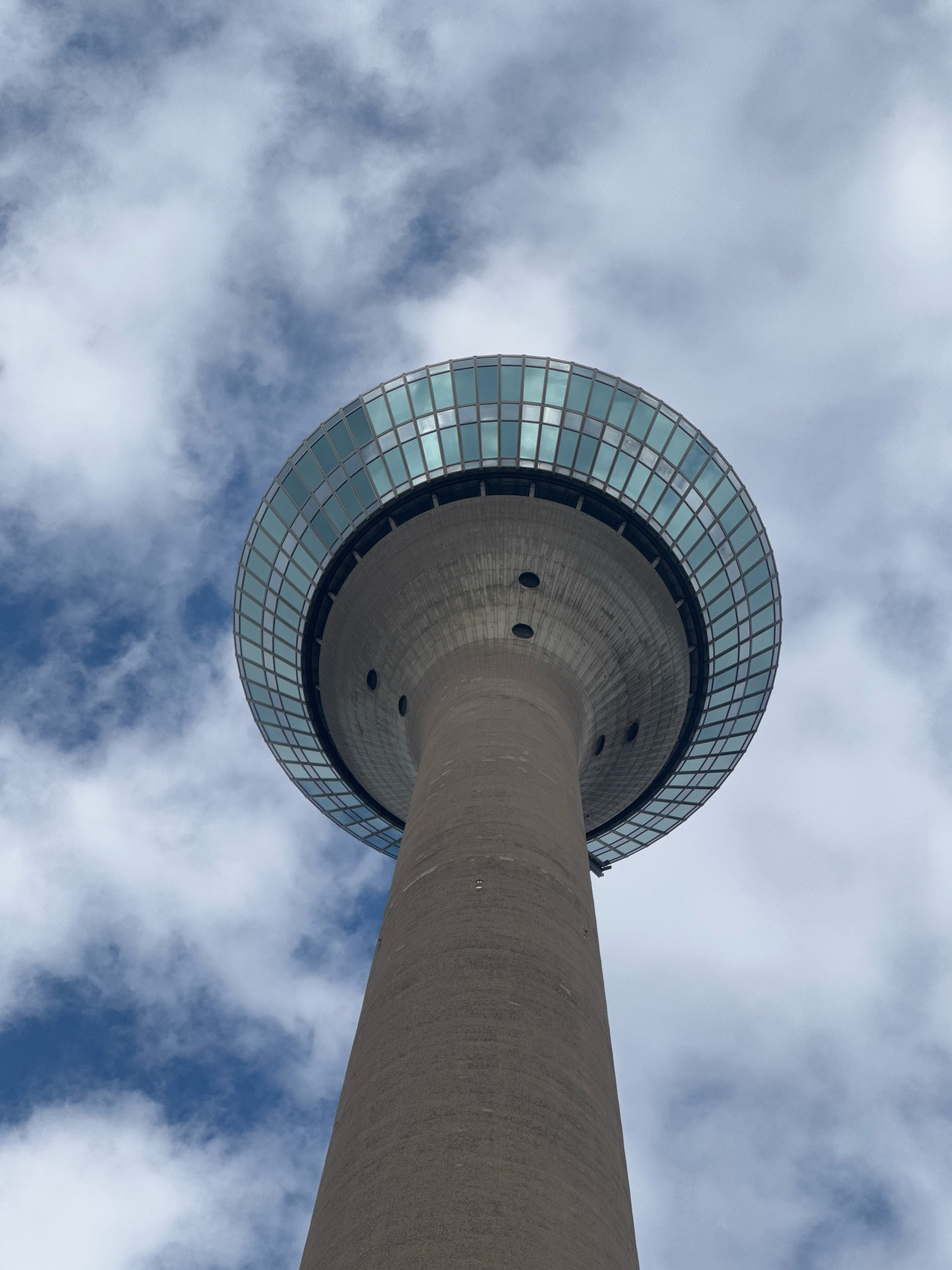 This is a low-angle, upward shot of the Rheinturm, a telecommunications tower in Dusseldorf, Germany, against a cloudy sky. The tower is made of concrete and has a circular observation deck with a glass facade at the top. The sky is filled with white and gray clouds, with patches of blue visible. The image is taken from below, emphasizing the height and scale of the tower.