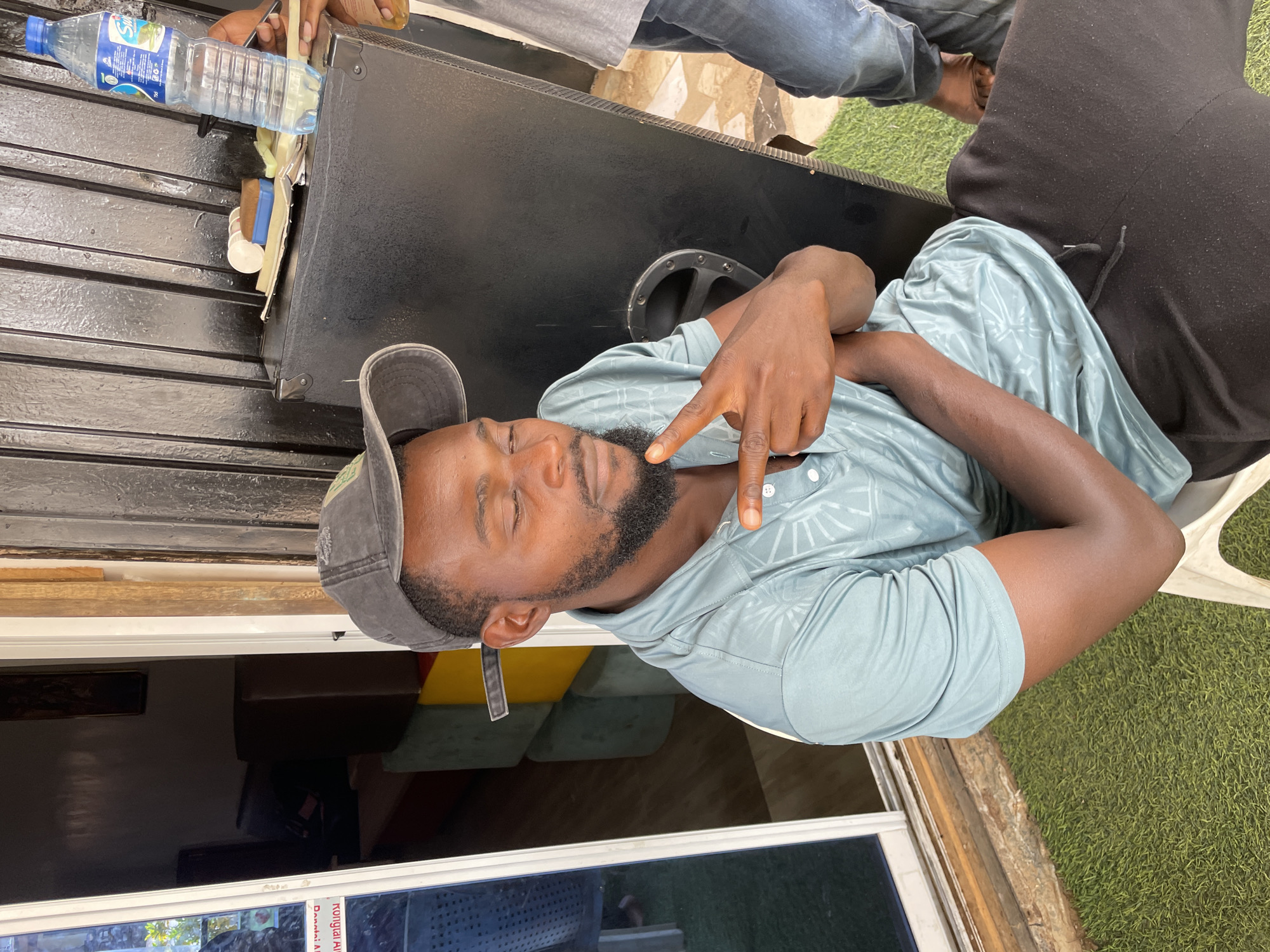 This image, set in Barakin Akawo, Nigeria, captures a casual, sunlit scene, likely outdoors or in a semi-open area. The primary subject is a man with dark skin and a beard, wearing a light blue short-sleeved shirt with a subtle pattern and a grey baseball cap reversed on his head. His eyes are closed, and he is making a "peace" or "victory" hand gesture with his right hand, suggesting a relaxed, confident, or playful mood as he poses for the camera.
He appears to be seated or leaning next to a large, black, rectangular object, possibly a speaker or sound system, which takes up a significant portion of the frame. On top of this black object, a clear plastic water bottle with a blue cap is visible, bearing the brand name "SWIFT" and "75 cl" on its label. Other small, indistinct items, including what looks like a white cup, are also on the surface.
Various parts of other individuals are visible around the main subject. Hands are seen near the water bottle in the upper left, while legs in blue jeans and bare feet are visible further up, suggesting another person is sitting on the black object. On the far right, part of a person wearing a black top is visible next to the main subject.
The background features green artificial turf or grass, indicating an outdoor or patio setting. Through a window in the lower left, faint glimpses of an interior space with furniture, possibly a brown couch and colorful cushions, can be seen. The bright, natural lighting suggests it is daytime.