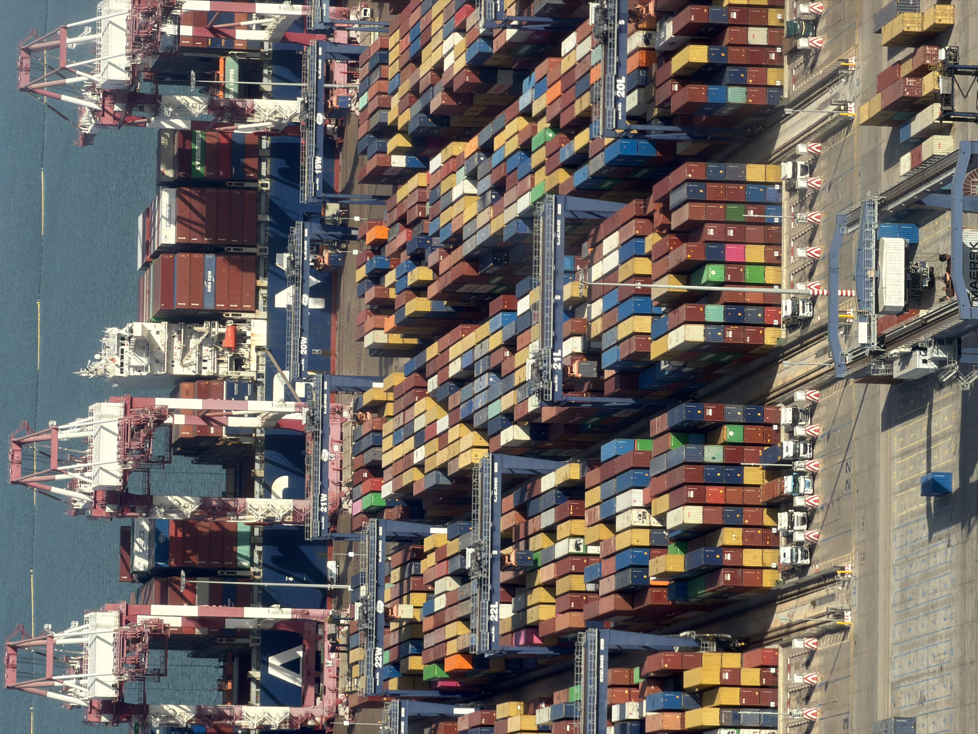 This is an aerial, high-angle photograph of a bustling container port, likely located in Barcelona, Spain, given the visual characteristics of the cranes and the types of cargo. The scene is dominated by massive, red and white gantry cranes that dwarf the numerous shipping containers stacked in intricate patterns. These containers, in a variety of colors including red, blue, yellow, green, and white, are organized in rows and columns, some reaching several stories high. They are being moved and organized by large port cranes and smaller vehicles, suggesting active cargo operations.
In the background, a large cargo ship is partially visible, laden with containers. The water of the port, likely the Mediterranean Sea, is a calm, deep blue. In the foreground, a paved area with markings indicates roads or operational zones, with several trucks parked and awaiting loading or unloading. One truck is actively engaged in moving a container under one of the gantry cranes. The overall impression is one of immense industrial activity and logistical efficiency. The lighting suggests it is daytime, with clear visibility and no significant atmospheric haze. The image appears to be a snapshot of a typical day at a major international shipping hub.