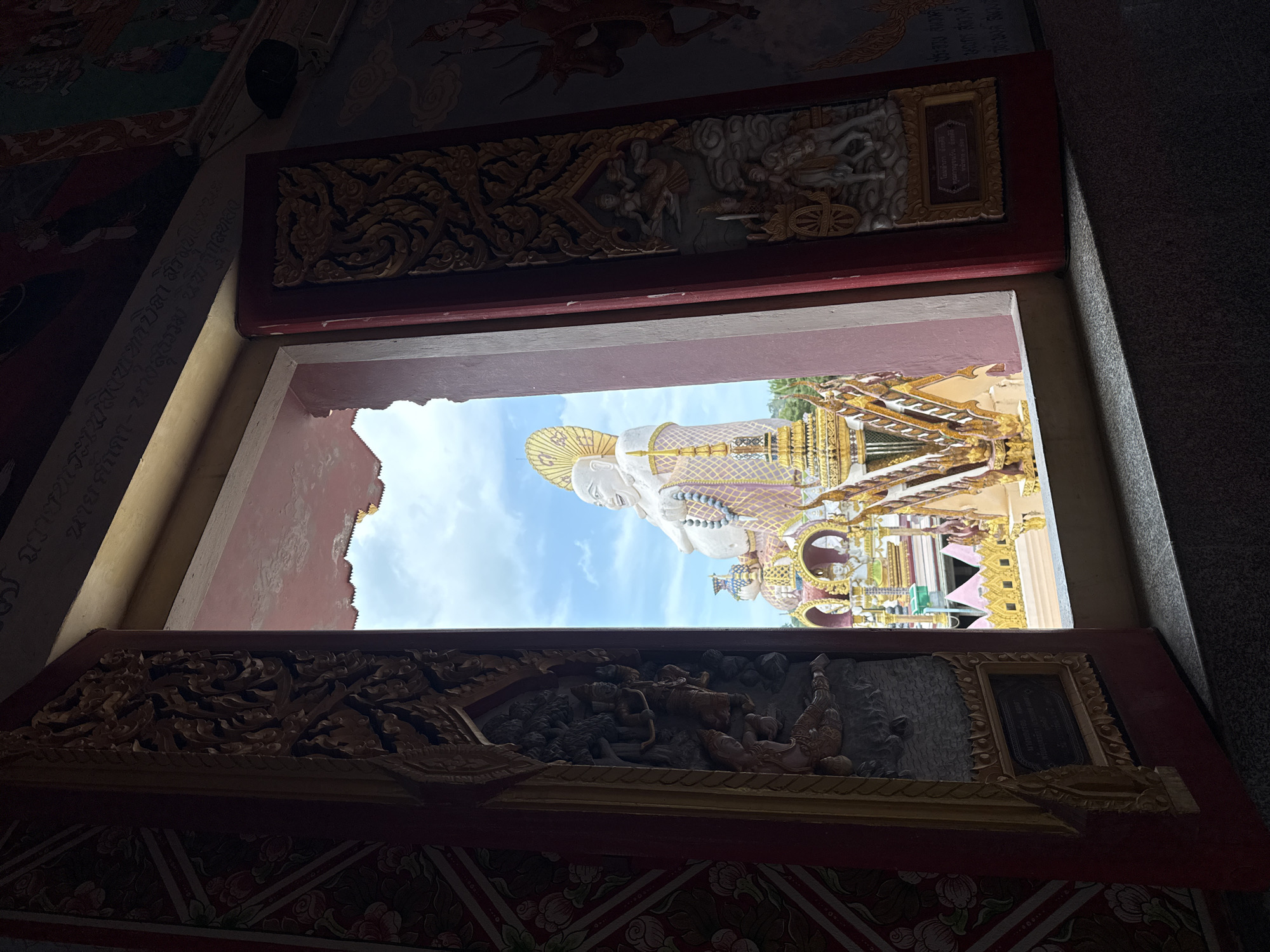 The image depicts a view through an open window, looking out onto a Buddhist temple complex in Ko Samui District, Thailand. In the foreground, the dark interior of a building is visible, with ornately carved wooden frames on either side of the window. These frames feature intricate patterns and bas-relief carvings of figures and mythical creatures. The window itself frames a bright, outdoor scene under a partly cloudy blue sky.
Dominating the outdoor view is a large, white, seated Buddha statue with a golden halo. The statue has a cheerful, rounded face and is adorned with a patterned robe. Below the Buddha statue, the temple's architecture is evident, characterized by golden spires and ornate, multi-tiered roofs, some of which are topped with decorative elements and statues. The colors of the temple structures are vibrant, with gold, pink, and white being prominent.
The walls to the right of the window opening are adorned with murals. On the far right, a mural depicts figures on horseback and mythological scenes. Closer to the window, a framed bas-relief carving is visible on the wooden structure, showing figures in a cart pulled by a horse, with other figures above. On the left side of the window, a section of a wall mural with floral patterns in shades of pink and green is visible. The overall impression is one of a rich cultural and religious site, seen from an interior vantage point.