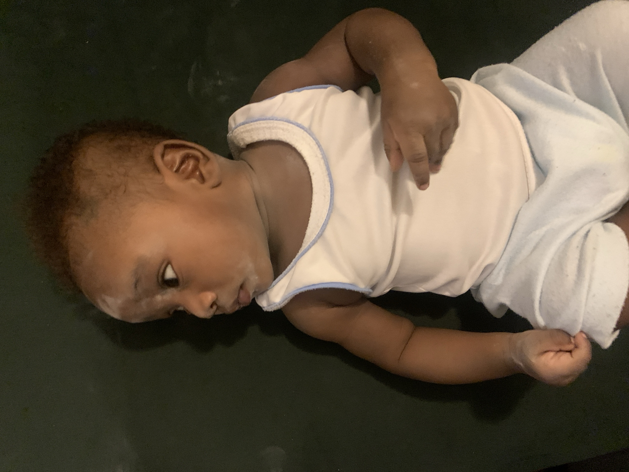 A baby lies on a dark surface. The baby wears a white shirt with a light blue trim and light blue pants. The baby's head is turned to the left. The location is unknown and there is no evidence to suggest that the image was taken in Barakin Akawo, Nigeria.