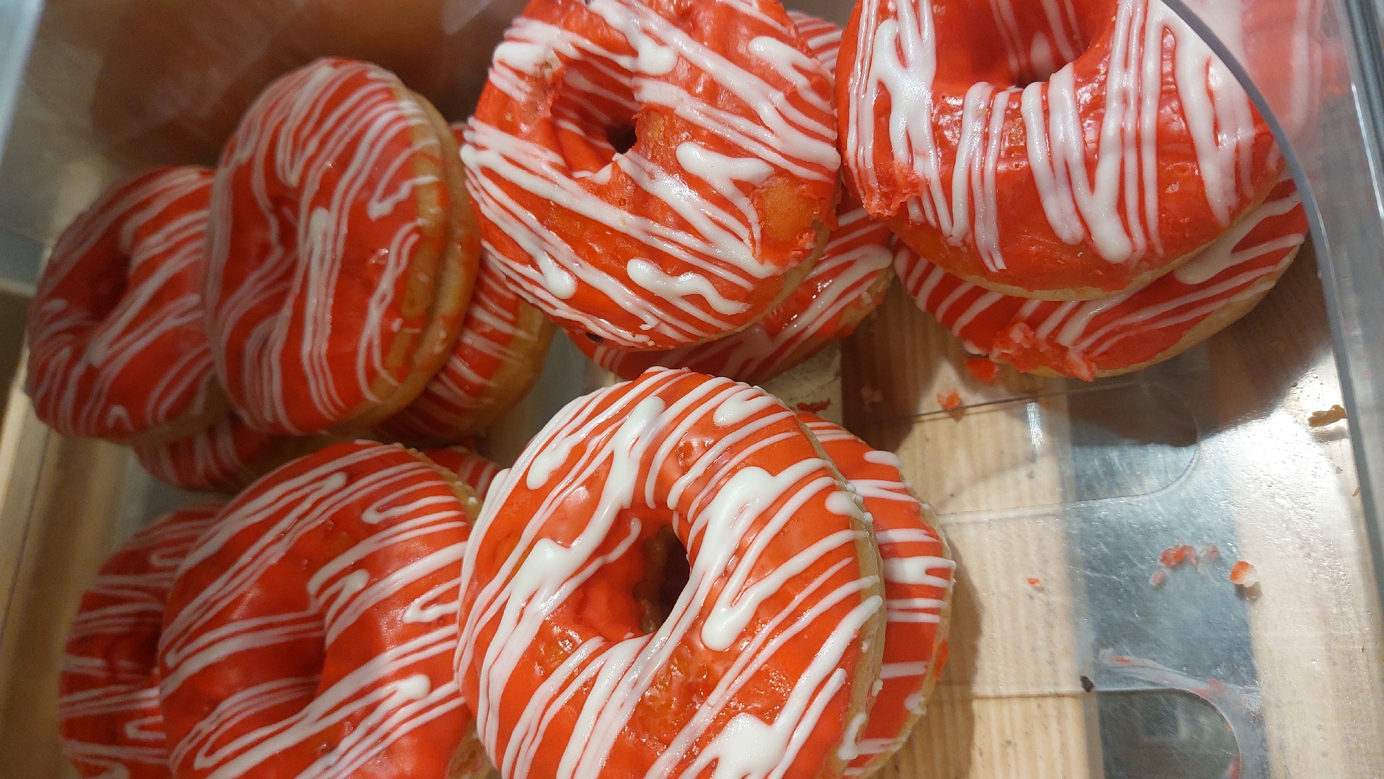 A top-down close-up shot shows a collection of ring donuts with bright orange frosting and white drizzle, arranged in a clear container. Some donuts are stacked. The setting appears to be a bakery or a food display case, based on the presentation. No specific location information, such as the city of Novosibirsk, Russia, is discernable from the image itself.