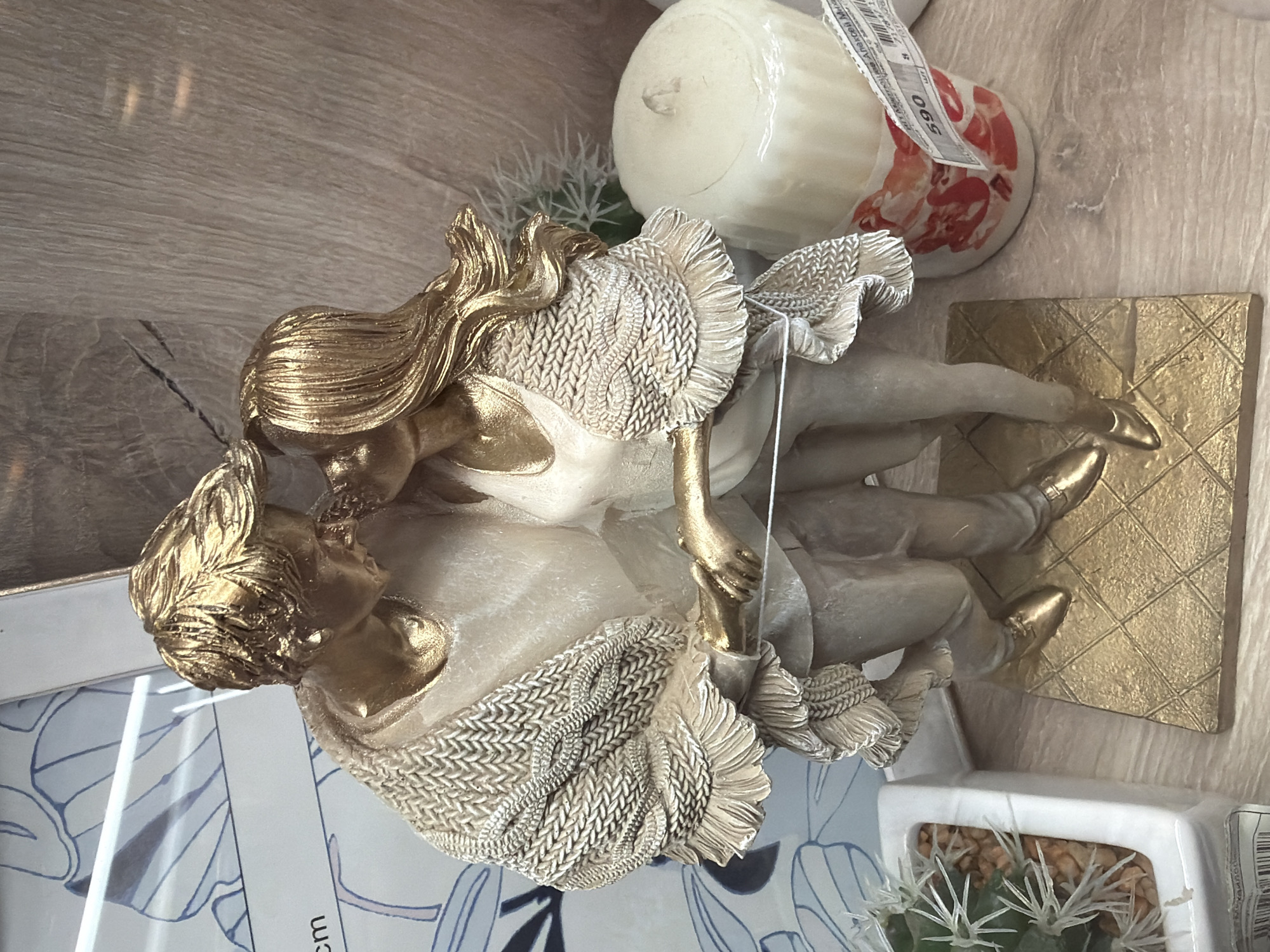 The image shows a kissing couple statue with golden accents, placed on a surface with various other items. To the right of the statue is a candle in a decorative holder. In the bottom right is a square, white planter with a small cactus. The statue rests partially on a box. The background surface appears to be a light wood grain. The location is Chaikovsky, Russia.