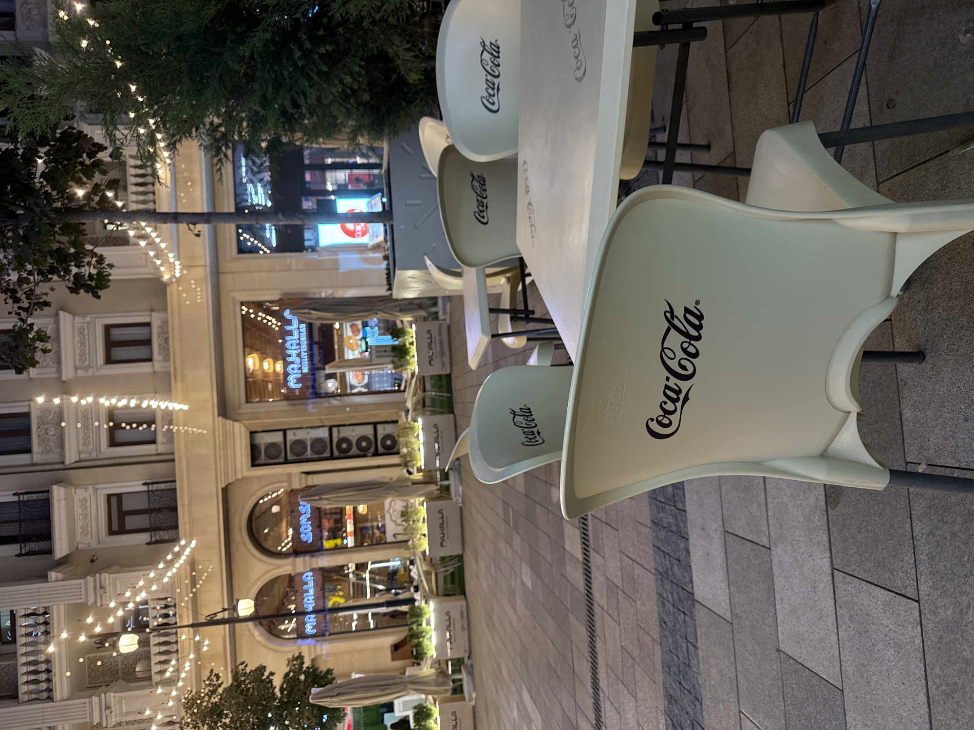 The image depicts an outdoor seating area of a cafe or restaurant at night, likely located in Tashkent, Uzbekistan, given the presence of the "Mahalla" signs visible on the building facade. The foreground is dominated by a light-colored table with several chairs around it, all featuring the Coca-Cola logo. The chairs are a pale cream color and have a curved backrest. The tables are also a similar light color.
In the background, a building with a classical architectural style is illuminated by strings of fairy lights, creating a festive atmosphere. Several arched entrances to shops or restaurants are visible on the ground floor, with signs such as "MAHALLA" and "SOMS" in Cyrillic and Latin script. The pavement in front of the building is paved with rectangular stone tiles. A large green coniferous tree stands to the right of the frame, further enhancing the outdoor setting. The overall impression is a pleasant evening scene in a public urban space.