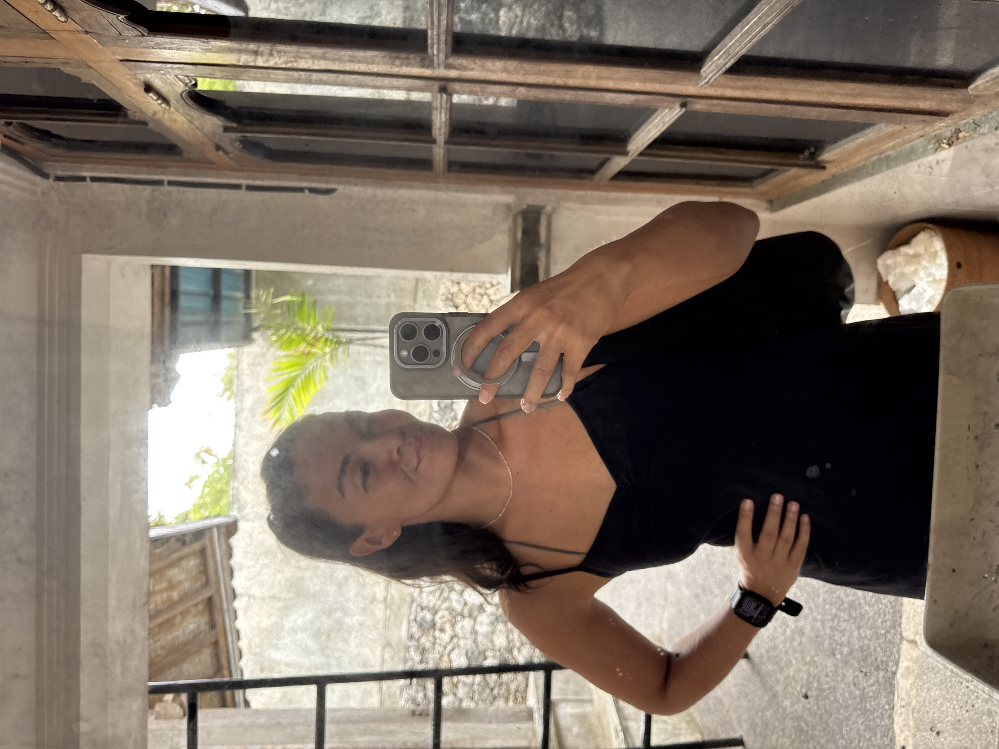 A woman takes a mirror selfie in a rustic, open-air setting. She is wearing a black slip dress and a thin silver necklace, and a black smartwatch on her left wrist. Her hair is dark brown and pulled back. The mirror reflects a stone wall, a wooden-framed window with arched panes, a metal ladder, and lush green foliage outside. The floor appears to be concrete or stone. A woven basket filled with white material is visible to the right of the woman, near the window. The overall ambiance suggests a tropical location, possibly a guesthouse or villa in Nusa Penida, Indonesia, given the architectural style and vegetation.