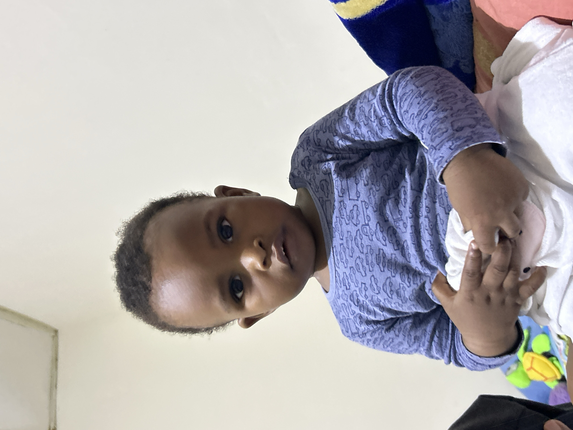 A young child, appearing to be around one to two years old, sits facing forward. The child has dark skin and short, curly black hair. They are wearing a long-sleeved, light blue shirt with a repeating pattern of cars. The child's hands are clasped together in front of them, holding a white blanket or piece of fabric. The background consists of a plain, light-colored wall. To the right of the child, a portion of a blue and yellow blanket or pillow is visible. In the lower left corner, a bright green and yellow turtle-shaped toy is partially visible, along with other colorful toys. The lighting suggests an indoor setting, possibly a bedroom. The overall impression is that of a domestic scene. The image does not provide any specific visual cues to indicate a location in Jos, Nigeria.