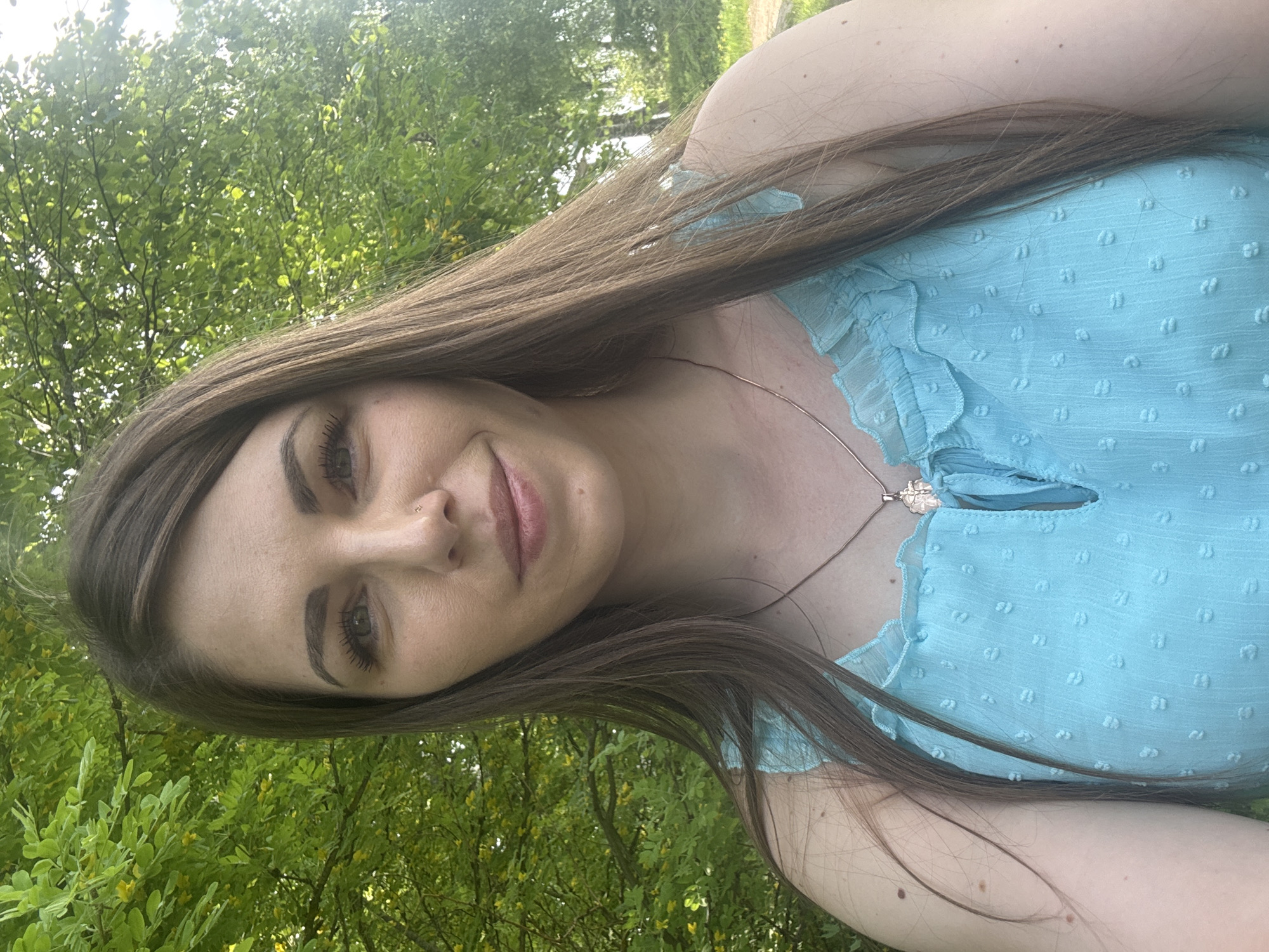 A woman with long, straight brown hair, fair skin, and brown eyes is photographed outdoors, possibly in a park or wooded area. She is wearing a light blue, textured top with ruffled details around the neckline. A delicate silver necklace with a pendant is visible. She is smiling faintly. The background consists of green foliage, suggesting a natural setting. The photo is taken from a slightly downward angle. The location is not possible to identify as Shevlyakovo, Russia based on the image.