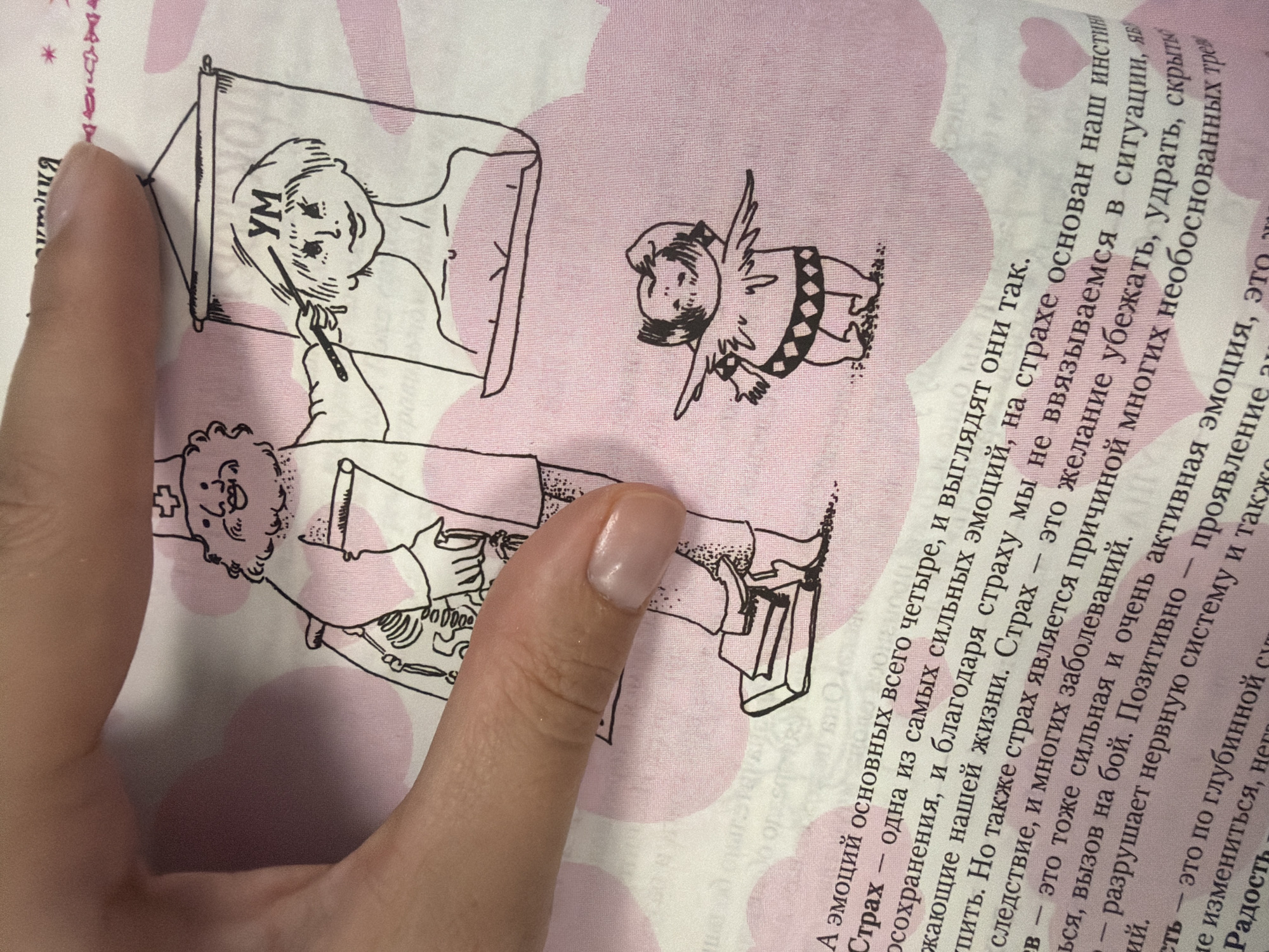 A hand with light skin and a natural-colored nail is holding open a book with a pink background and a repeating pattern of light pink circles. The book contains a cartoon illustration and text. The illustration depicts a doctor-like figure with a beard and hat pointing a wand at a framed portrait of a woman's head, on which the letters "УМ" are written on the forehead. A small winged creature stands to the right of the doctor-like figure. The text is in Russian and discusses emotions such as fear. The scene is indoors, with the lighting suggesting it is daytime. There is no discernible connection to the city of Homyel, Belarus.