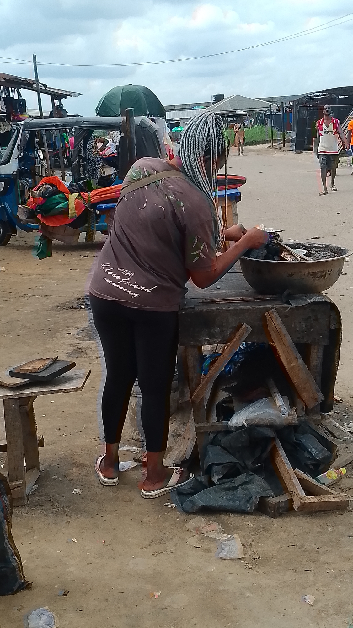 A person with numerous thin, light-colored braids extending from their head stands behind a wooden table in an outdoor market in Warri, Nigeria. The person is wearing a loose-fitting brown t-shirt with an emblem on the back that reads "2023 Close friend recurrence" and black leggings, with white and pink sandals on their feet. They are actively engaged in sorting charcoal from a large, shallow metal bowl on the table. The table itself is a rustic wooden structure, with some items like bags and wood scraps piled beneath it. To the left of the table, a smaller wooden stand holds two flat, dark objects, possibly wood or stone. In the background, there are several market stalls with corrugated metal roofs, various goods displayed, and other people walking around. A blue and white auto-rickshaw, also known as a "keke," is partially visible on the far left, laden with colorful textiles and other items. The ground is dirt and appears somewhat dusty, with scattered debris. The sky is overcast.