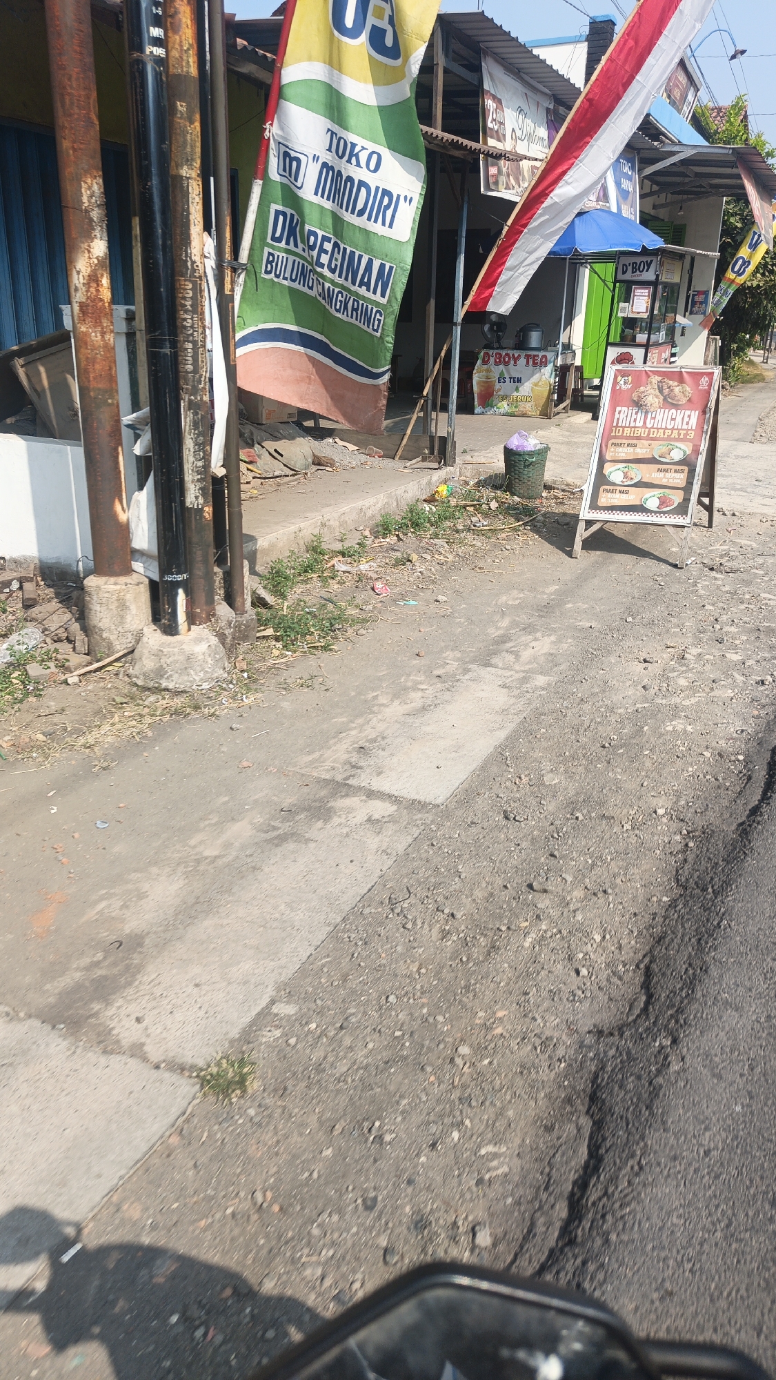 The image shows a street scene in Bulungcangkring, Indonesia, with a focus on a small shop or food stall. A tall, green banner with text that appears to be in Indonesian, including "TOKO 'MANDIRI'" and "DK PECINAN BULUNG SANGKRING," is prominently displayed, hanging from a pole. To the right of this banner, a red and white Indonesian flag is also visible. In front of the shop, a "D'BOY TEA" sign and a "FRIED CHICKEN" advertisement are placed on the sidewalk, along with a green trash can. The ground is paved with concrete and asphalt, showing some wear and tear. Vegetation is present along the edges of the pavement, and debris is scattered in some areas. The scene suggests a commercial area, possibly a roadside eatery or market. The perspective suggests the photo was taken from a moving vehicle, likely a motorcycle, given the presence of handlebars and a mirror in the foreground.
