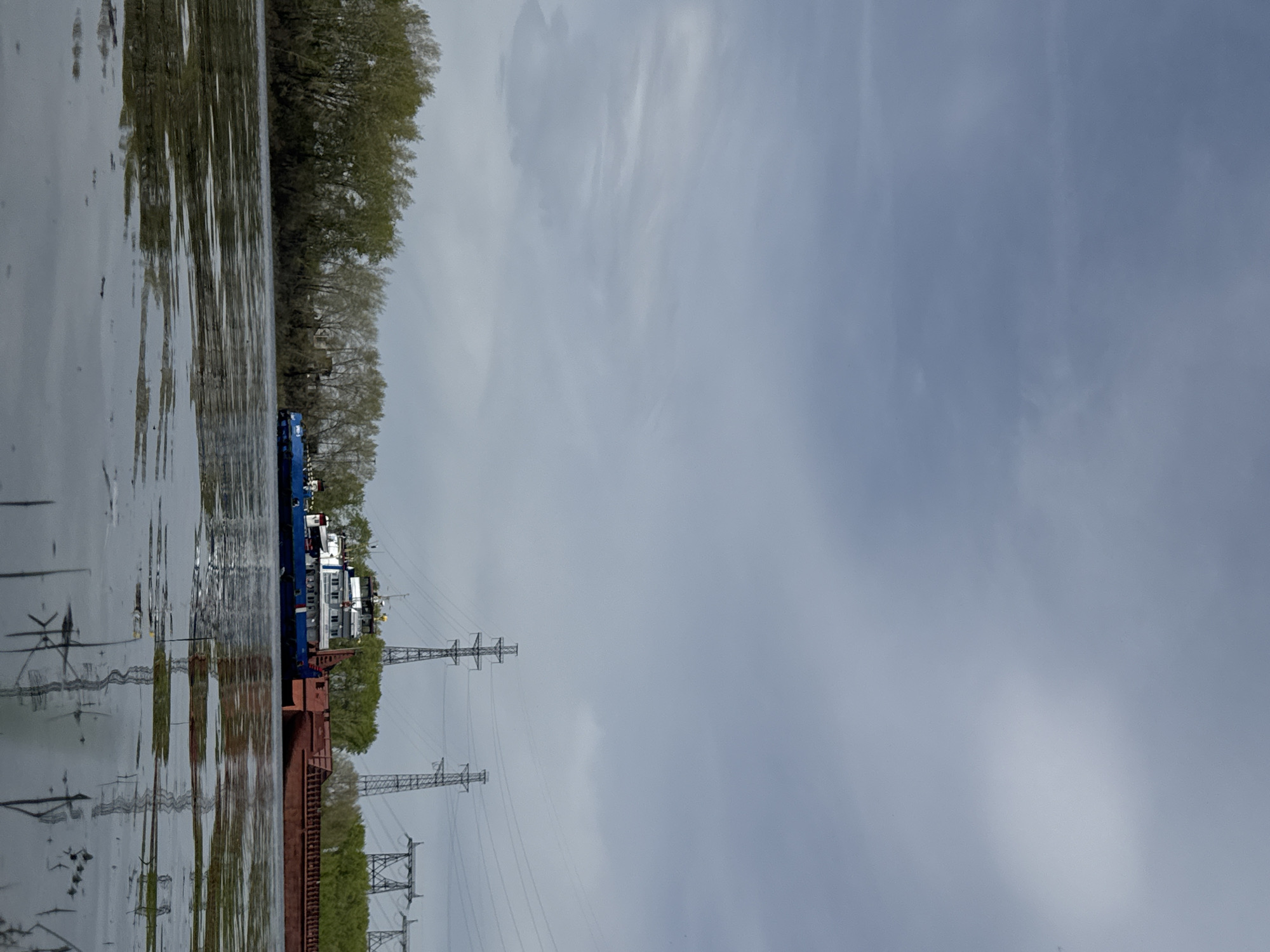 The image depicts a scene on the Kama River in Chaikovsky, Russia. A large, blue and white barge is docked or moving slowly along the river's edge. A rusty, brown barge is connected to it. Power lines and pylons are visible on the riverbank, running parallel to the river. The opposite bank is lined with trees, which are reflected in the water. The sky is overcast with grey clouds. There are no people visible in the shot.