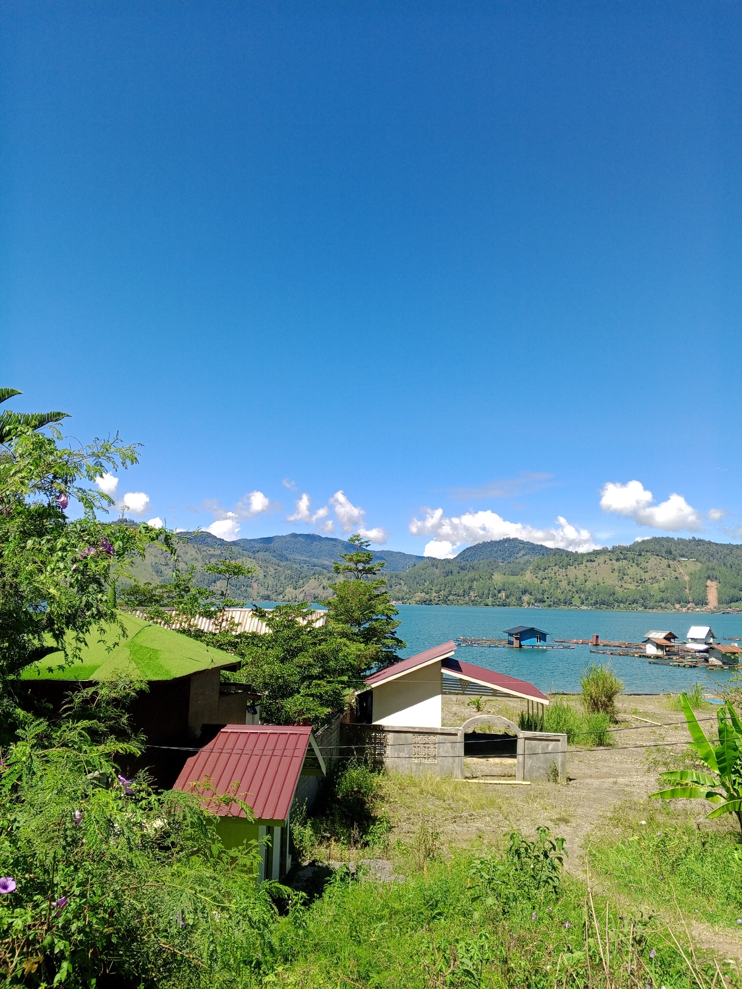 This is a landscape photograph of a body of water with floating structures, taken on a clear day. The foreground features lush green vegetation and several small buildings with red and green roofs. A light-colored building with a red-tiled roof is partially visible on the right side, with a concrete structure in front of it. Further back, a vibrant blue lake stretches out, dotted with various floating structures that appear to be houses or platforms. Some of these structures have dark blue or white roofs. The lake is surrounded by rolling hills covered in dense green trees and foliage, and in the distance, there are mountains under a bright blue sky with scattered white clouds. The scene suggests a tranquil, possibly rural, environment. The image is captured from a slightly elevated vantage point, looking out towards the lake and mountains.