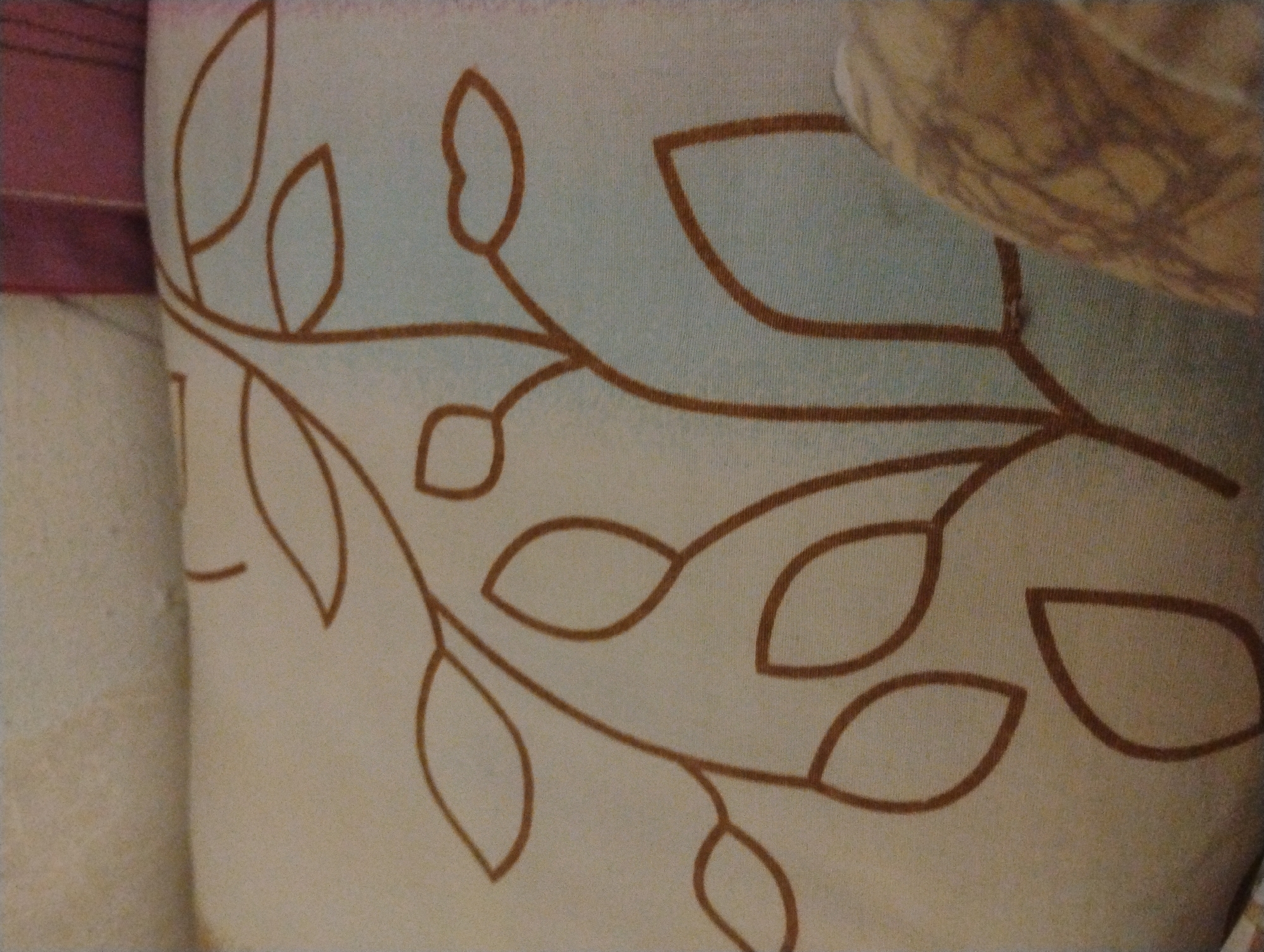 The image displays a close-up, low-angle shot of a cream-colored fabric cushion with a brown, stylized floral or leafy pattern. The pattern consists of thin, curved brown lines forming abstract shapes that resemble leaves and stems. The cushion appears to be soft and possibly textured, as suggested by the subtle variations in light and shadow on its surface. In the lower right corner, a portion of another object, possibly a piece of furniture or another cushion, is visible. This object has a pattern of brown and beige, appearing somewhat textured and organic. The background is out of focus and consists of muted colors, suggesting indoor lighting. There are no people or discernible actions taking place in the image. The overall impression is one of a domestic setting, likely a living room or bedroom, with a focus on decorative soft furnishings. There is no information within the image that provides any context related to Plachkovtsi, Bulgaria.