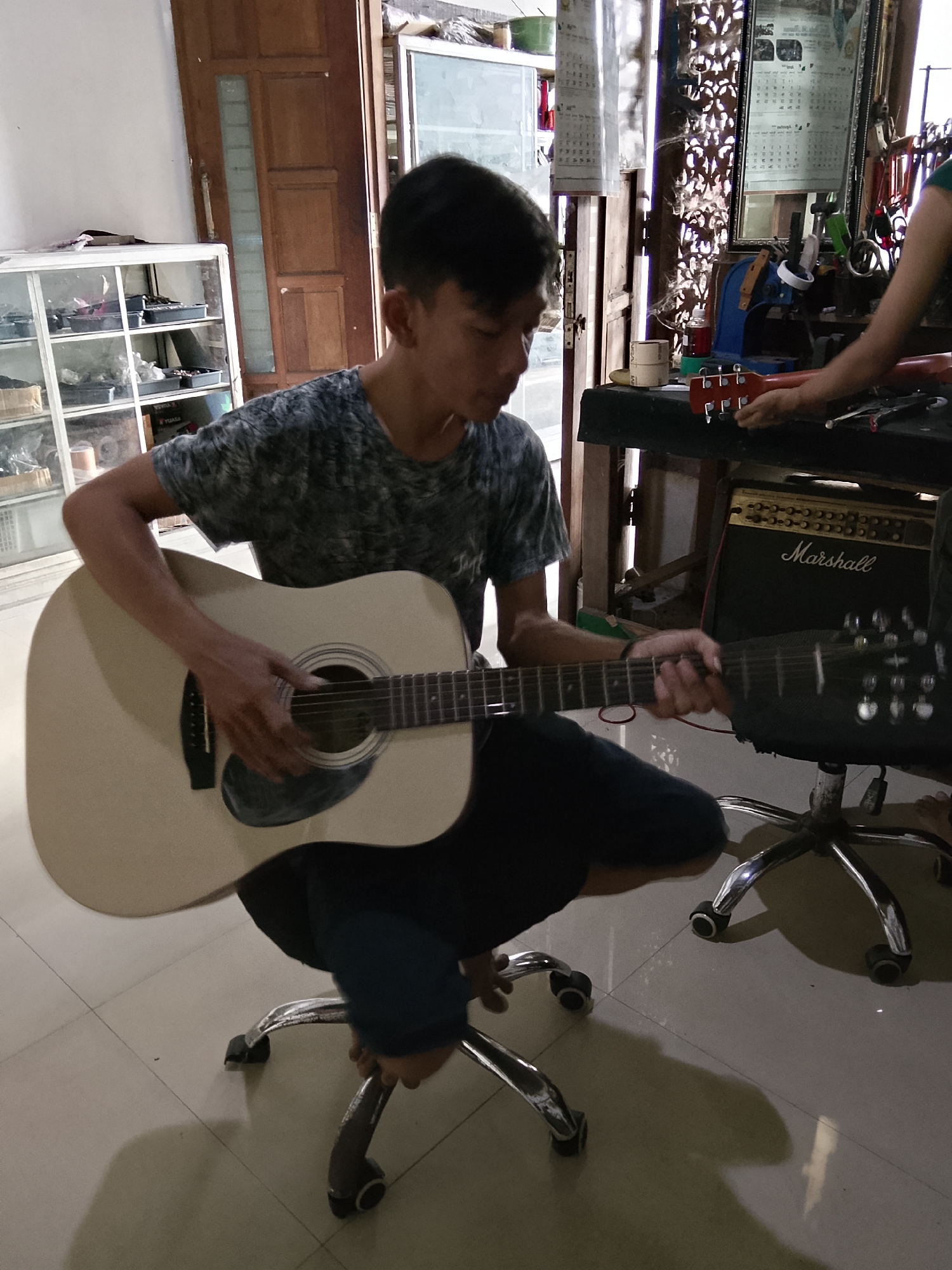 In Dahanrejo, Indonesia, a young Indonesian male is depicted seated on a rolling office chair, actively playing an acoustic guitar. The guitar is light-colored with a dark pickguard and visible tuning pegs. He wears a dark t-shirt with a gray pattern and dark trousers. The scene unfolds in what appears to be a workshop or storage area, evidenced by shelves filled with various items in the background, including what seem to be boxes and containers. A wooden door with glass panes is visible to the left, and a Marshall amplifier is positioned behind the guitar player. Another person's hands are seen reaching towards a guitar neck from the right side of the frame, suggesting a collaborative or instructional setting. The floor is tiled, and artificial lighting casts shadows.