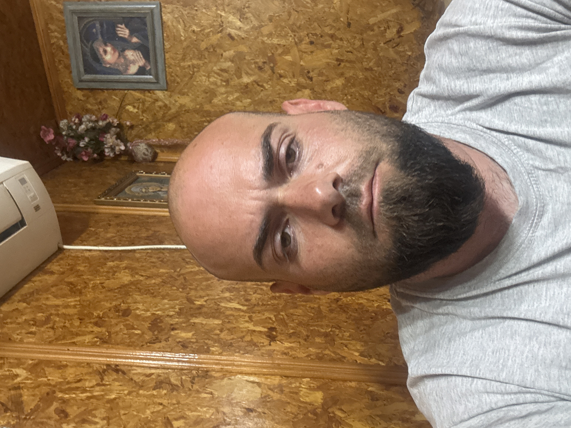 A selfie-style photograph shows a bald man with a dark beard wearing a gray t-shirt. The background appears to be a wood-paneled wall. Mounted on the wall are a framed picture of people, a small bouquet of artificial flowers, and a framed picture of religious imagery. The left side of a white air conditioning unit is partially visible. The location cannot be verified with the information provided.