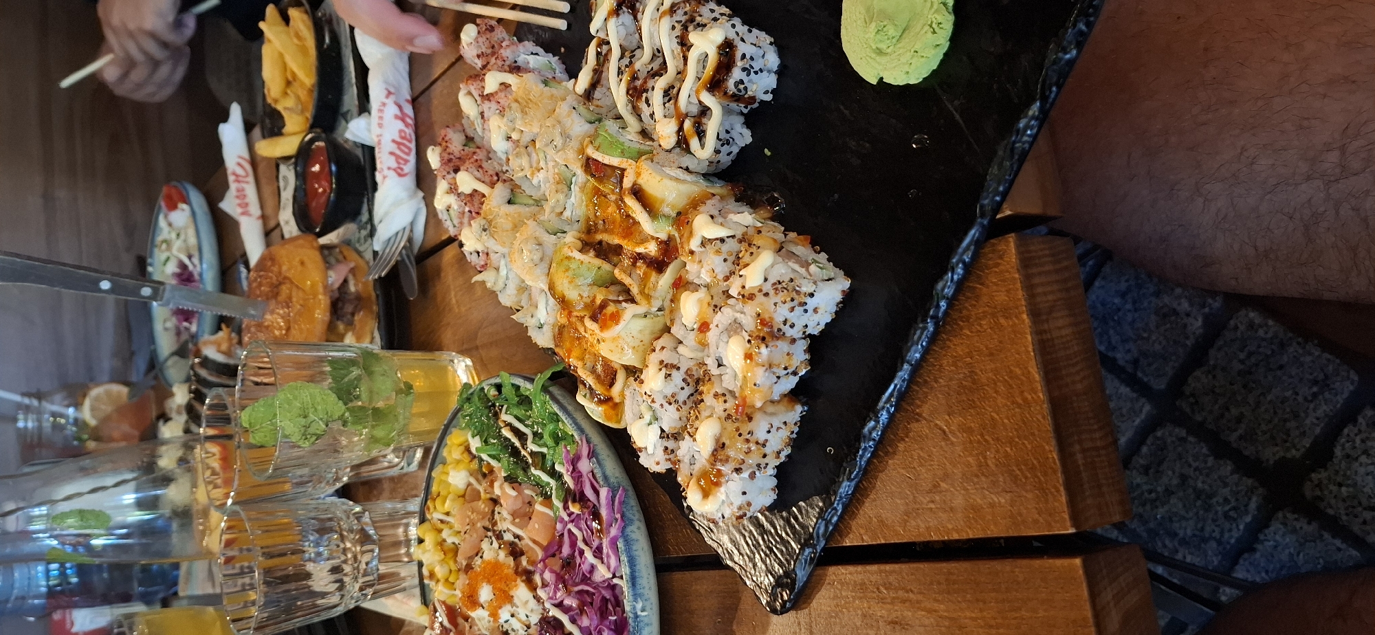 The image depicts a table laden with food and drinks, suggesting a meal is in progress at a restaurant. The food items include a burger impaled with a knife, a bowl of poke or salad with corn, red cabbage, and what appears to be salmon, a plate of various sushi rolls, and a side of french fries. Beverages in glasses, some with mint, are also visible. The setting appears to be an indoor dining area with wooden furniture and a patterned rug visible beneath the table. The overall impression is one of a casual dining experience.
Without additional information or context, it is impossible to determine the specific location in Burgas, Bulgaria.