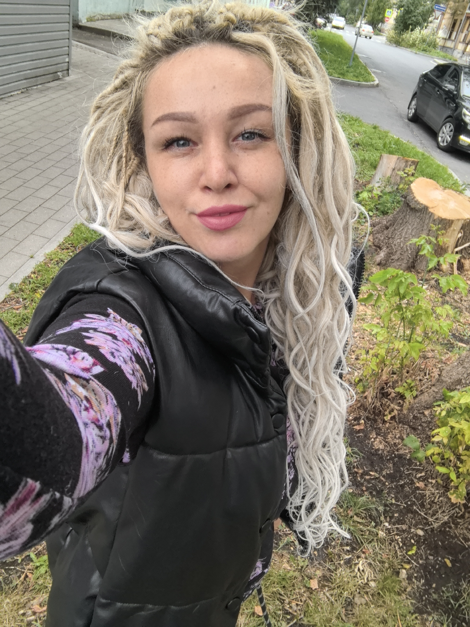 A woman with blonde dreadlocks takes a selfie outdoors. She is wearing a black puffer vest over a black long-sleeved shirt with a purple floral pattern. She is also wearing pink lipstick. The background shows a sidewalk, a street with cars, and some trees and foliage. The image was taken in Nizhny Novgorod, Russia.