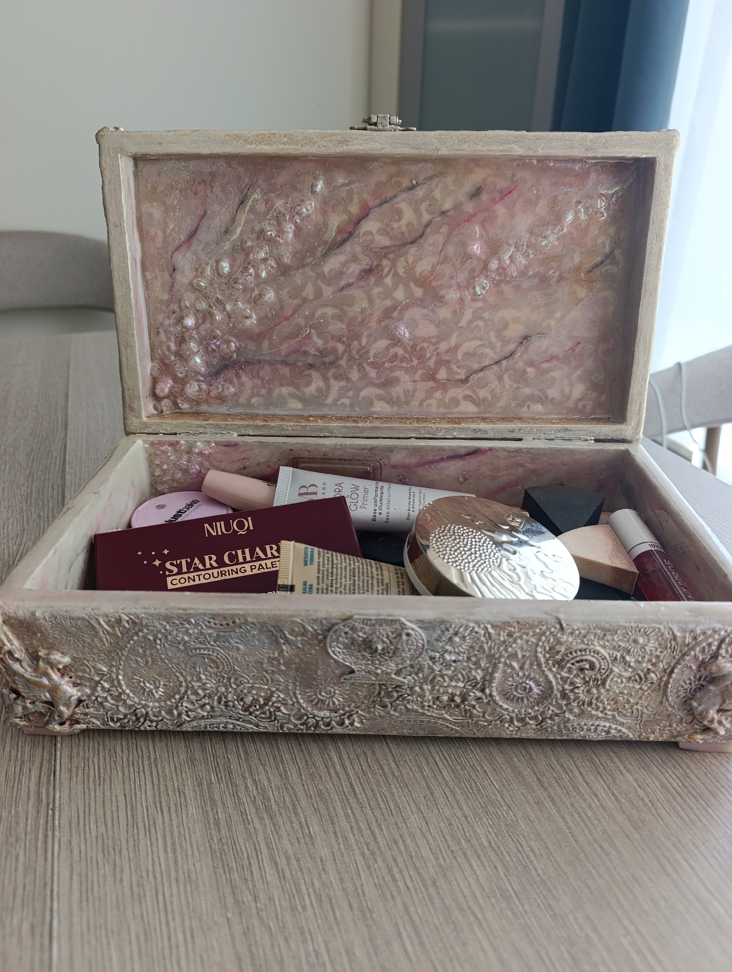 An open, ornate wooden jewelry box sits on a light-colored wooden table. The interior of the box is lined with a pink and white marbled resin with pearlized accents. Inside the box, a variety of makeup products are neatly arranged. These include a burgundy eyeshadow palette labeled "NIUQI STAR CHARMING CONTOURING PALETTE," a small pink compact, a tube of "B. GLOW PRIMER," a silver-colored compact with a textured lid, a dark gray triangular sponge, a light beige wedge-shaped sponge, and a clear tube with a red liquid, likely lip gloss, labeled "STUBBORNLY." The exterior of the box is decorated with raised, intricate paisley patterns in an antique white finish, with some darker accents adding depth. The box is positioned in the foreground of the image. The background shows a blurred view of a room with furniture, suggesting an indoor setting. The location is not Toruń, Poland.