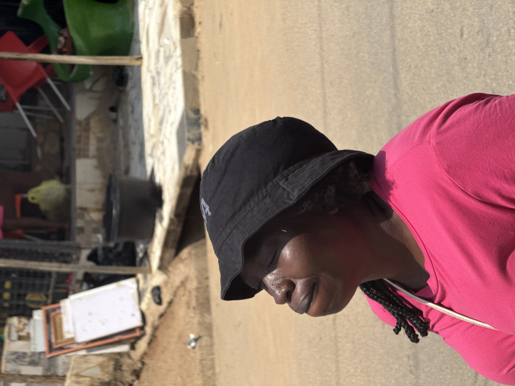 A medium shot captures a dark-skinned woman wearing a black bucket hat and a bright pink t-shirt. Her eyes are closed, and her face is tilted upwards, seemingly basking in sunlight. Sweat glistens on her forehead and cheeks, suggesting a warm, possibly humid, environment. Her lips are slightly parted, and her expression conveys a sense of peace or contentment. One braid of her dark hair is visible over her left shoulder. A thin white strap runs diagonally across her chest, possibly from a bag or clothing.
The background is blurred but suggests an outdoor urban setting. To the left, there are stacked white and orange rectangular objects, possibly frames or boards. Behind these, dark plastic crates and other indeterminate items are visible. A large black bucket or tub is positioned in the mid-ground, and further back, red chairs and a green object, possibly a large container, can be discerned. The ground is a sandy or dusty surface, consistent with the dry, arid climate often found in parts of Nigeria. The lighting indicates it is daytime, with harsh sunlight creating strong highlights on the woman's face.
While the image does not explicitly state its location, the description aligns with typical street scenes found in urban areas of Abuja, Nigeria, which experience a tropical savanna climate with hot, dry seasons. The presence of street vendors, various recycled materials, and everyday objects in the background are common elements of such an environment.