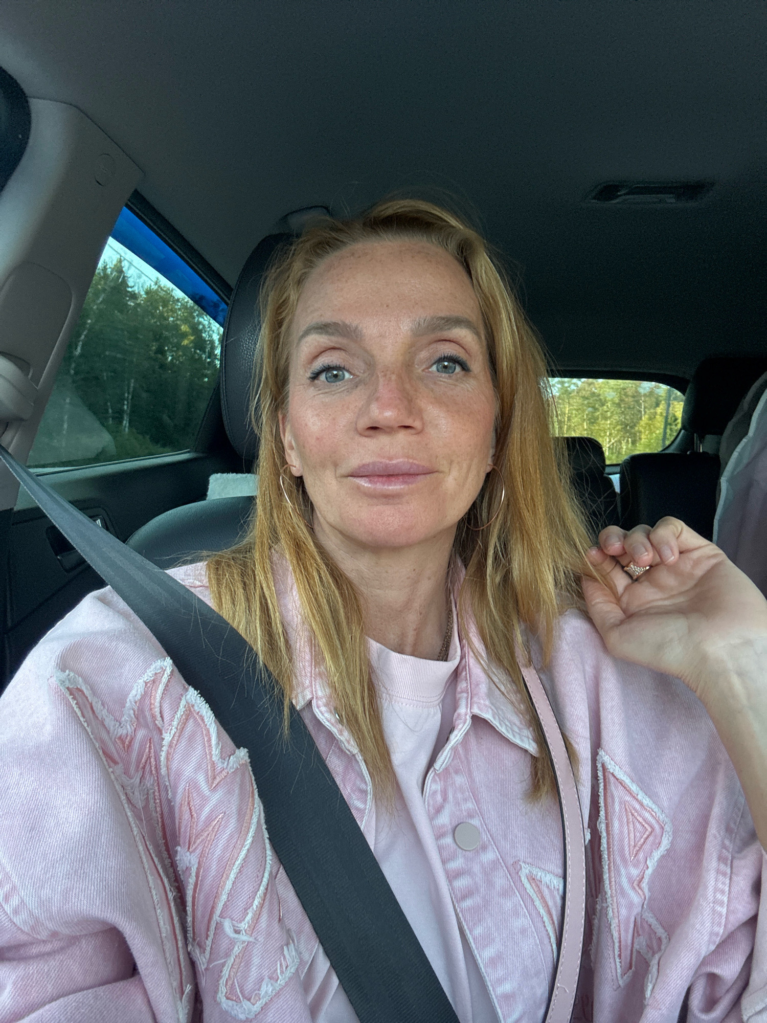 A blonde woman with freckles sits in the driver's seat of a car, wearing a light pink denim jacket over a pink t-shirt. She has blue eyes and is looking at the camera, with her left hand touching her hair near her face. The seatbelt is fastened across her body. The background shows the interior of the car, including the dashboard, a car window with trees visible outside, and part of a black headrest. The lighting suggests it is daytime. The location is not specified within the city of Unknown, Russia.