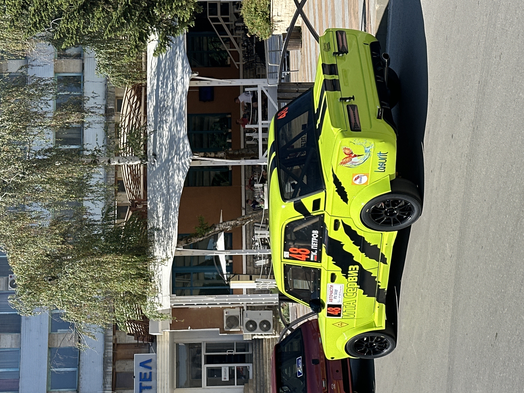 The image captures a vibrant, neon-yellow racing car parked on an asphalt surface in what appears to be a public area. The car, a modified sedan, is adorned with black racing stripes, the number "48" prominently displayed on its doors and roof, and various sponsor decals. Visible text on the car includes "ВИТА Сервиз", "АВТОЧАСТИ", "PIT STOP", and "К. ПЕТРОВ", suggesting it's associated with a racing team or event. A small sticker with a bird graphic and the word "Lasurit" is also visible on the rear quarter panel.
In the background, a building with a light-colored facade and multiple windows is present. A canopy-like structure extends from the building, providing shade over an outdoor seating area with tables and chairs, where several individuals are seated. Trees with lush green foliage are strategically placed around the scene, adding a natural element to the urban setting. The car is positioned on the right side of the frame, angled slightly towards the viewer, with a portion of a dark red car visible to its left. The lighting suggests it is daytime, with bright sunlight casting shadows. Based on the text visible on the car, specifically "ВИТА Сервиз" which is a known entity in Gorna Oryahovitsa, Bulgaria, it is highly probable this photograph was taken in the city of Gorna Oryahovitsa, Bulgaria.