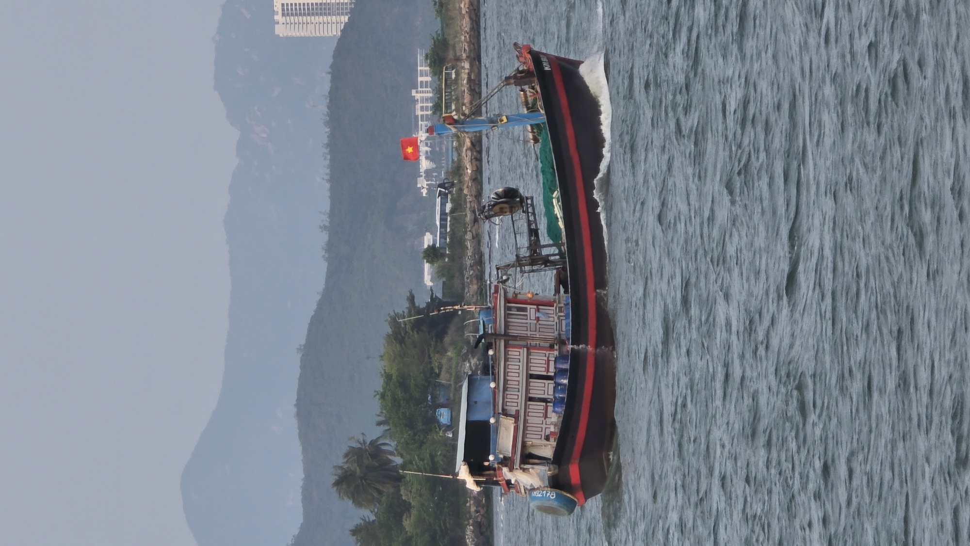 The image captures a fishing boat moving through the waters off the coast of Nha Trang, Vietnam. The boat, characterized by its dark hull with a red stripe, bears the inscription "Khanh Hoa," indicating its origin. A Vietnamese flag is prominently displayed on the boat, signifying its national identity.
In the background, the cityscape of Nha Trang is visible. Mountains rise in the distance, partially obscured by haze. The sea itself is choppy, with whitecaps indicating some wind action. The boat is leaving a wake, confirming its forward motion.