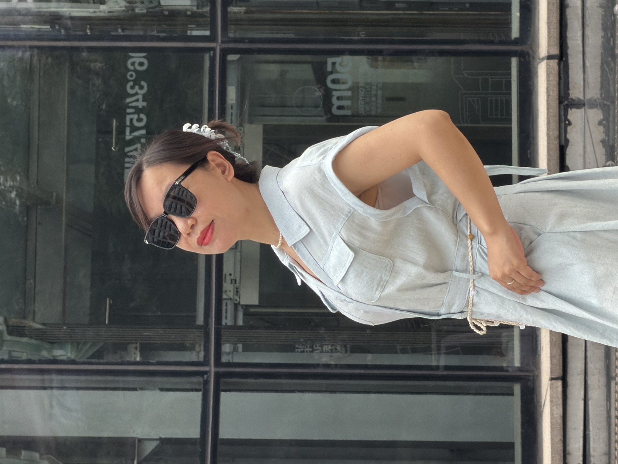 A woman stands outdoors in front of a glass building. She has dark hair pulled back with a white clip and wears large, dark sunglasses and a pearl necklace. She is dressed in a light blue collared dress with short sleeves, a belt made of braided cord, and a ring on her right hand. Her head is tilted back, and her lips are painted red. The background shows reflections in the glass of buildings and signage, including what appears to be text in Chinese. The building's architecture suggests an urban setting.