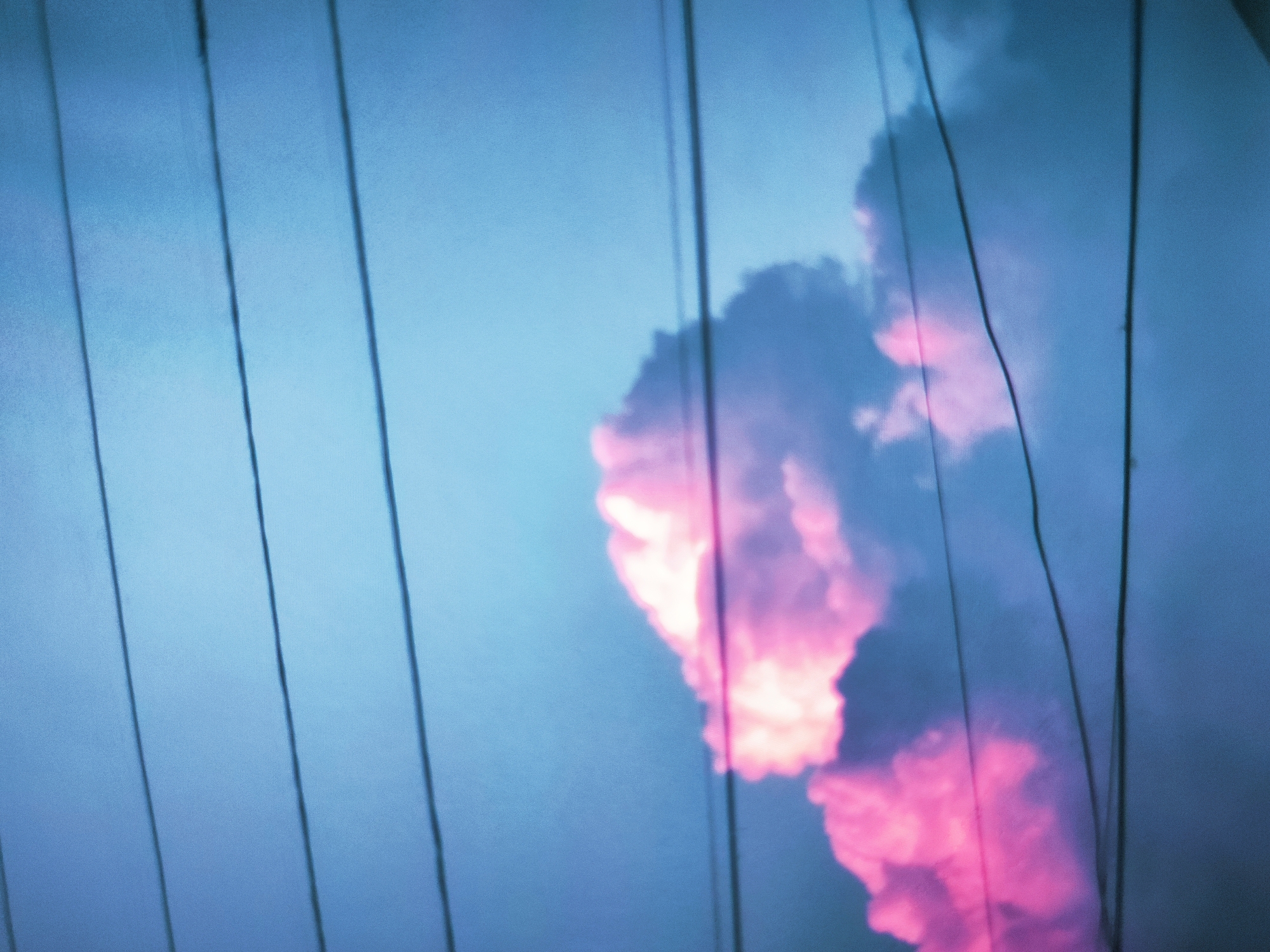 The image is an abstract, low-angle shot of a cloudy sky. The sky is predominantly a pale blue with streaks of bright pink and white, indicating sunset or sunrise. Several dark, thin, vertical lines, possibly power lines or wires, cut across the frame, partially obscuring the sky and clouds. The overall effect is impressionistic, with a focus on color and light rather than distinct forms. No people, buildings, or identifiable landmarks are visible. The image is from Bökönbaev, Kyrgyzstan.