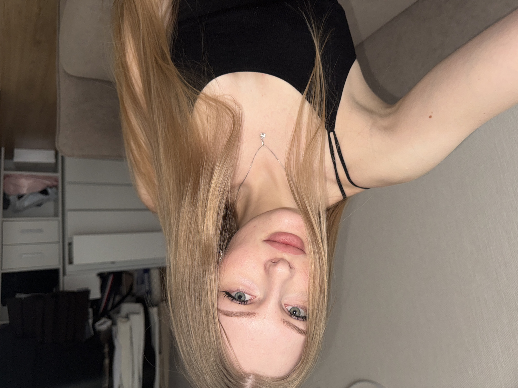 A young woman with long blonde hair, fair skin, and blue eyes is pictured. She is wearing a black tank top with thin straps and a delicate silver necklace with a pendant. The image is taken from an unusual angle, appearing to be upside down from the viewer's perspective. Behind the woman, there is a wardrobe with white drawers and hanging clothes visible. A neutral-colored sofa or couch is also partially visible in the background. There is no indication of this image being taken in Perm, Russia.