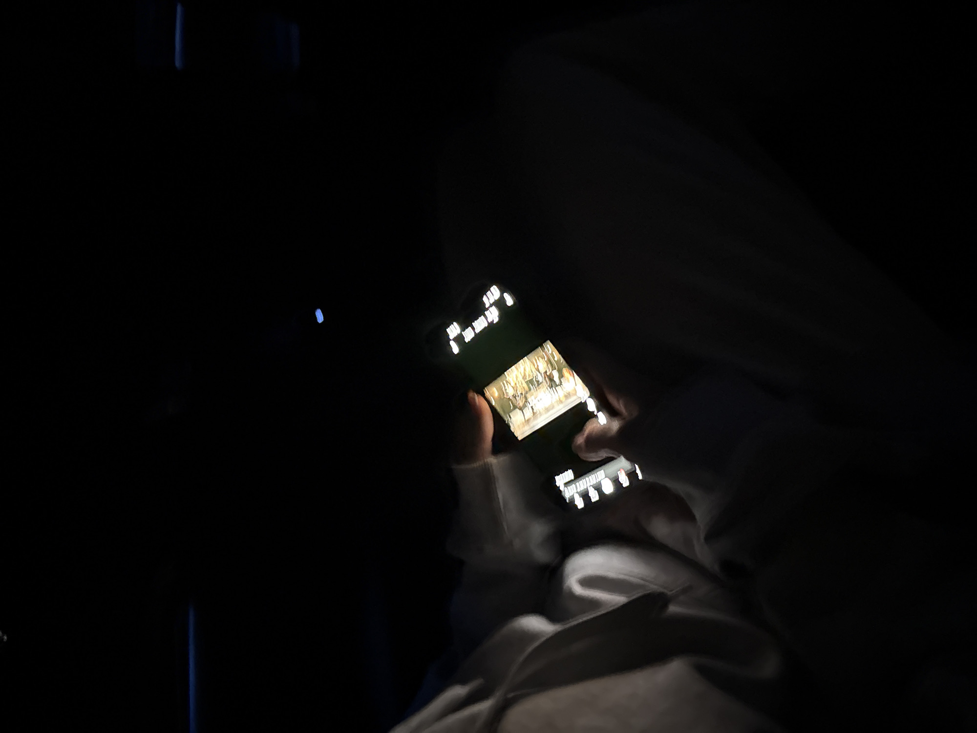 A person is holding a phone in a dark environment, possibly a car. The phone screen is illuminated, displaying what appears to be a video or image. The person's hands are visible, and they seem to be interacting with the phone. The environment is mostly dark, with some faint shapes suggesting the presence of a seat and possibly some interior features of a vehicle. The location is not identifiable as Kuchak, Armenia from the given image.