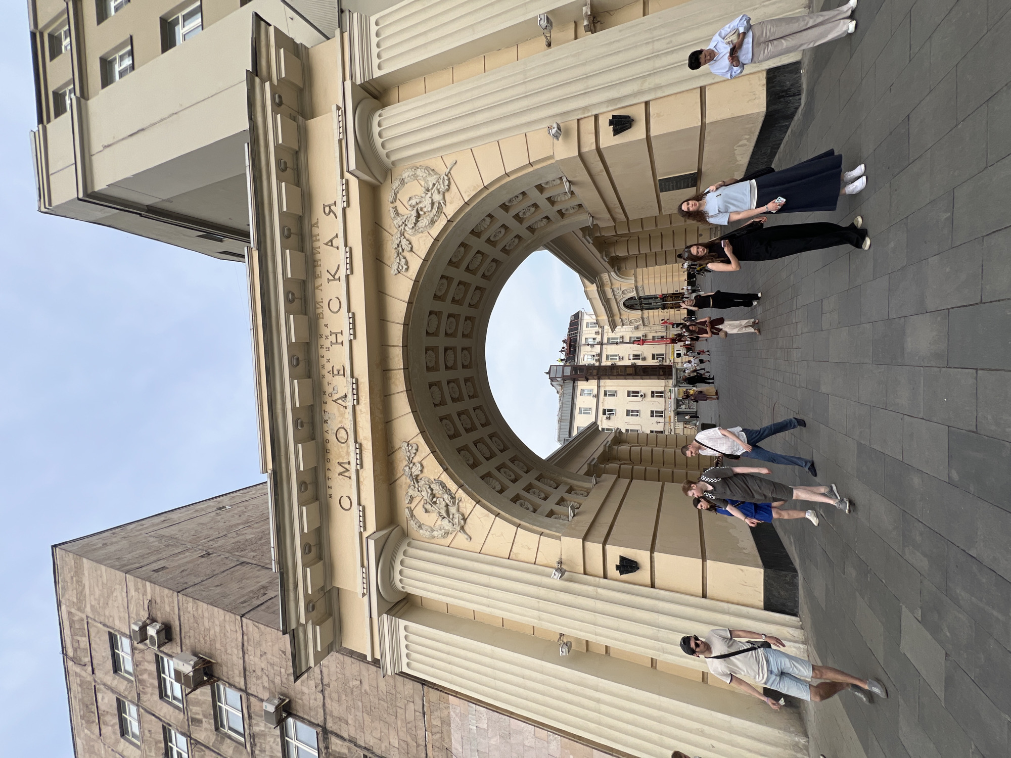 The image captures the entrance to Smolenskaya metro station in Moscow, Russia. The station's grand archway, adorned with classical columns and inscriptions, stands prominently. The inscription on the arch reads "МЕТРОПОЛИТЕН ИМЕНИ В.И. ЛЕНИНА" (Lenin Metro) and "СМОЛЕНСКАЯ" (Smolenskaya). A crowd of people is seen walking through the archway and on the pavement. The architecture suggests a historical Soviet era design. The sky is overcast.