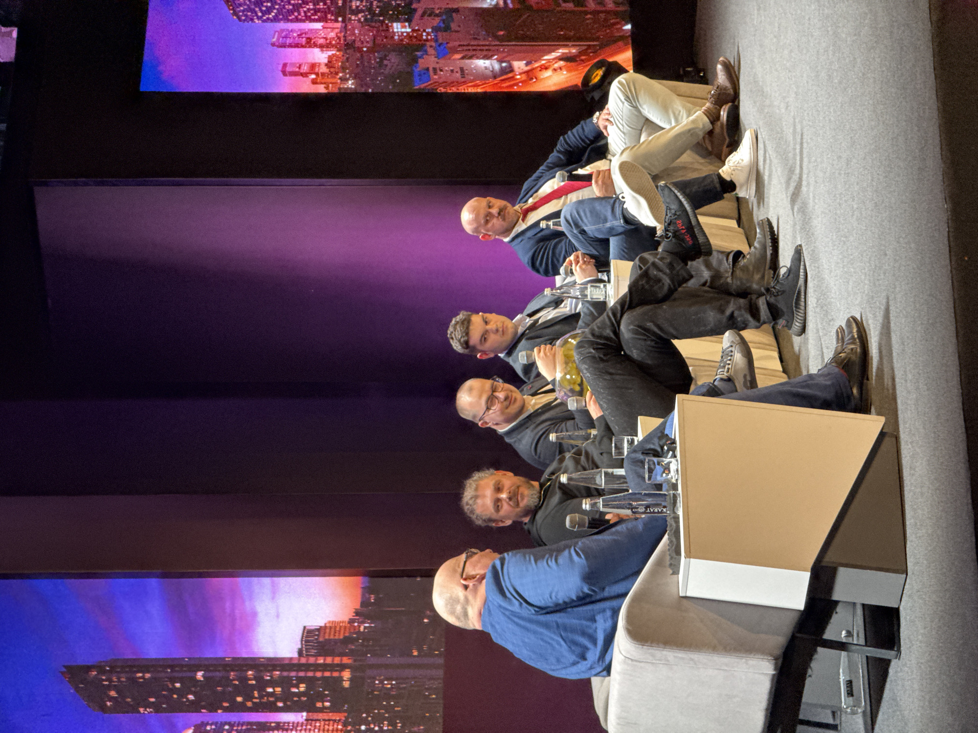 Five men are seated on a stage, likely during a panel discussion or conference. The backdrop features images of a cityscape. The men are dressed in business casual attire with water bottles on a table in front of them. Microphones are also present, suggesting they are speaking or preparing to speak. The event appears to be taking place in Perm, Russia, given the "KARAT" water bottles on the table, which are bottled in Perm. The specific location within Perm is not discernible from the image.