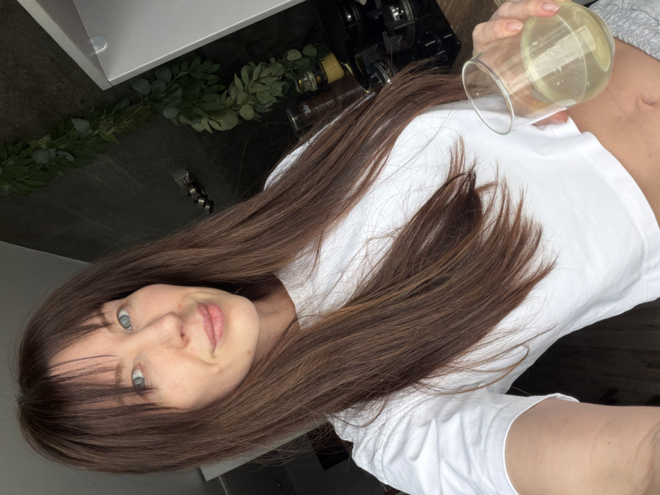 The image depicts a female individual taking a selfie indoors, within a kitchen located in the city of Perm, Russia. The woman, appearing to be in her 20s or 30s, has long, straight brown hair with bangs and blue eyes. She is looking directly at the camera with a slight smile. She is attired in a white, short-sleeved, cropped t-shirt, revealing her midriff, which exhibits a faint mark near the navel. Her left hand is holding a stemless clear glass containing a pale yellow liquid. The background reveals a contemporary kitchen environment featuring dark-colored tiled walls, a partial light-colored upper cabinet on the right, and a dark countertop. A black gas hob with four burners is visible on the countertop, accompanied by several glass jars. A green leafy decorative garland is affixed to the dark wall behind the subject.