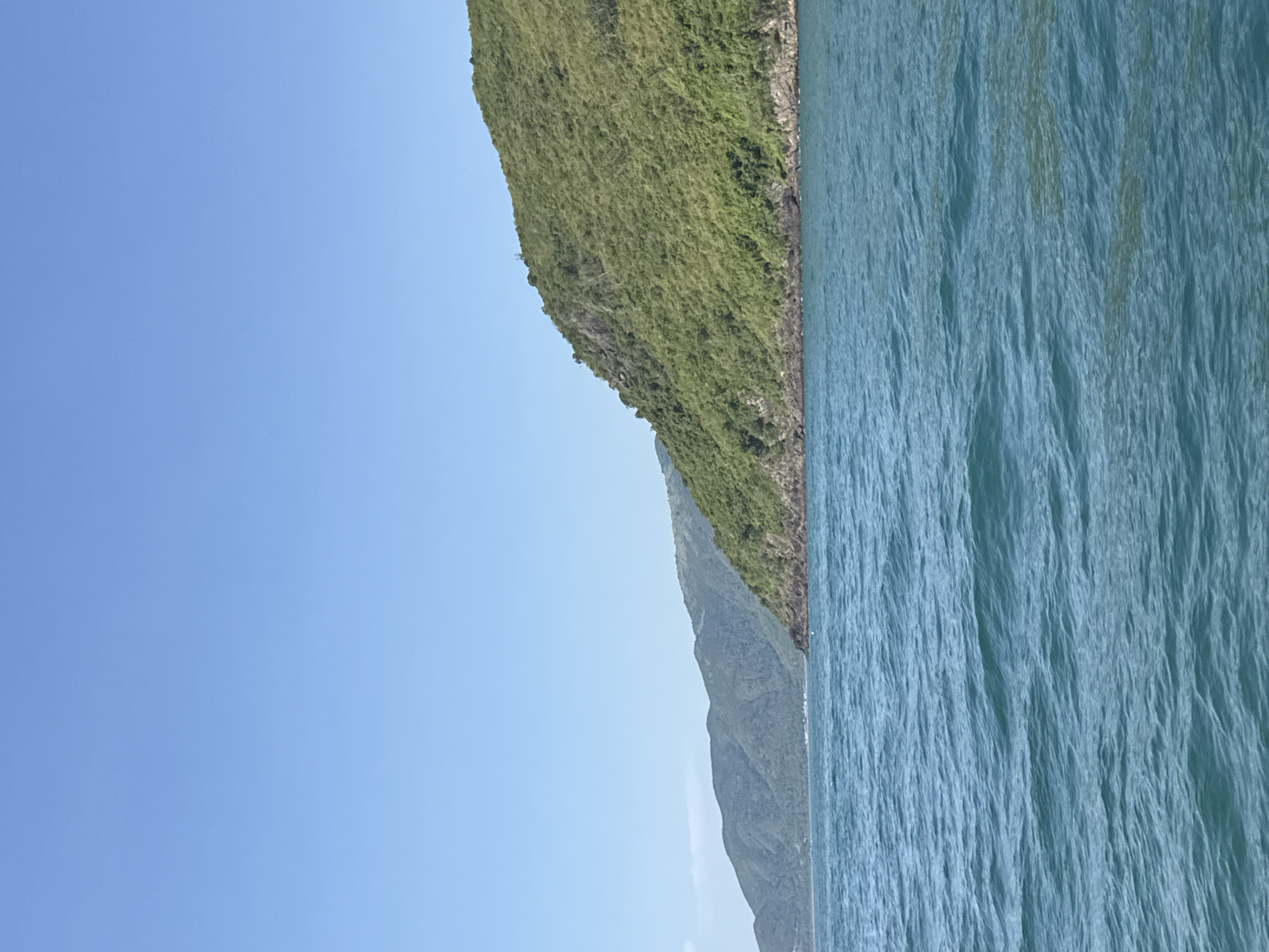 A wide-angle, eye-level shot captures a tranquil seascape against a backdrop of lush, green mountains under a clear blue sky. The foreground is dominated by the calm, turquoise waters of the ocean, with gentle ripples suggesting a slight breeze. The middle ground features the coastline, where the water meets rocky outcrops and the steep, verdant slopes of the mountains. These mountains rise majestically, covered in dense foliage, indicative of tropical vegetation. In the distance, a further range of hazy, blue-green mountains stretches across the horizon. The scene is devoid of any man-made structures or human presence, presenting a pristine, natural landscape. The lighting suggests it is daytime, with the sun likely high in the sky, casting even illumination across the scene. The image evokes a sense of peace and natural beauty, characteristic of coastal Vietnam, likely in a less developed or protected area.