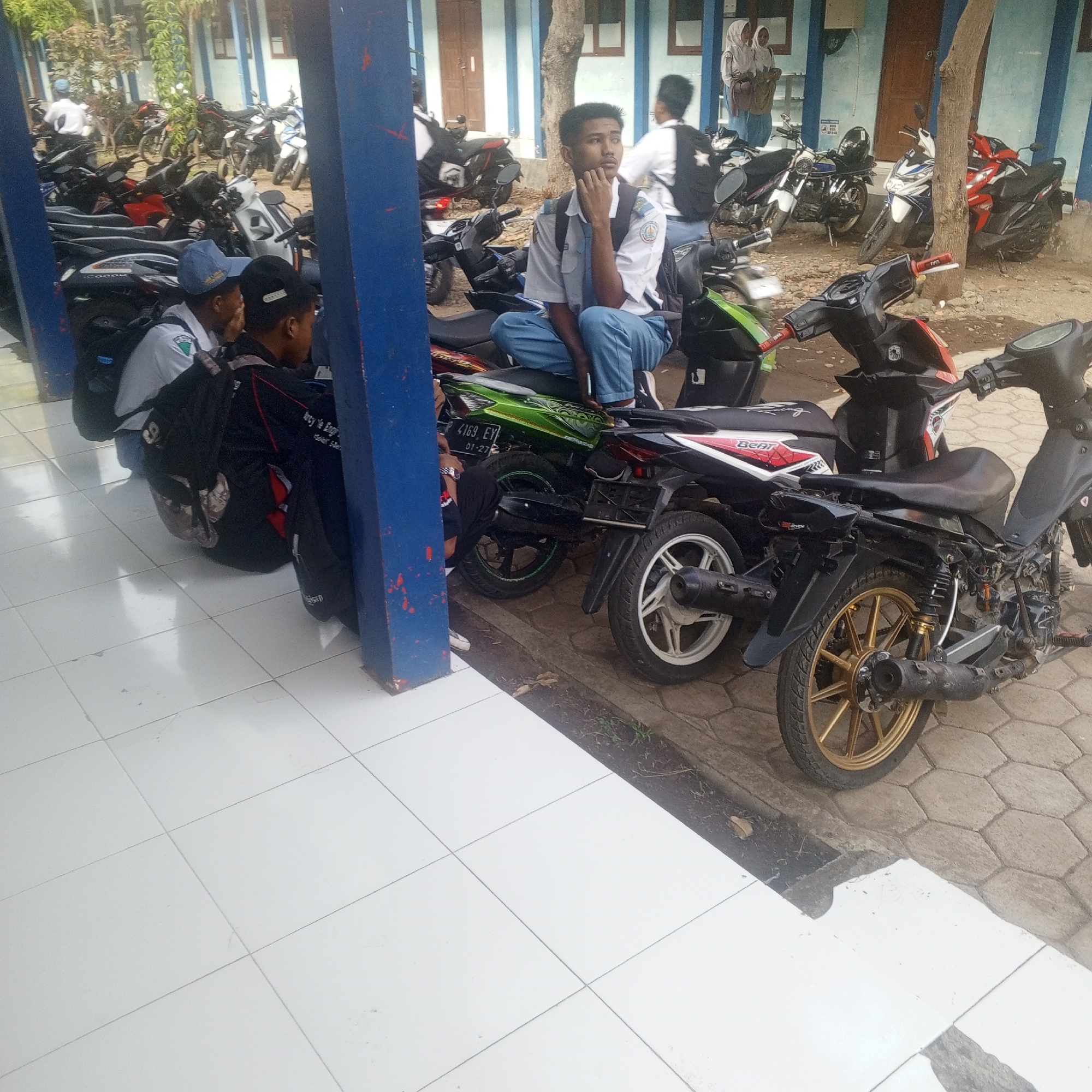 In Buduan, Indonesia, outside a building with blue and white walls and large windows, a group of young people in school uniforms are gathered around parked motorcycles. The foreground is dominated by a tiled walkway and a large blue column. Several students are seated on or near motorcycles, with some looking at their phones or talking amongst themselves. One student in the center is sitting on a green motorcycle, wearing a white shirt with a blue collar and tie, and blue pants, with a backpack on. Another student to his left, wearing a blue cap and white shirt, is partially obscured. Several motorcycles of various colors, including red, black, and white, are parked in a lot behind the students. The ground is a mix of pavement and dirt. The overall scene appears to be a typical school day in Indonesia, with students waiting or socializing around their mode of transportation.