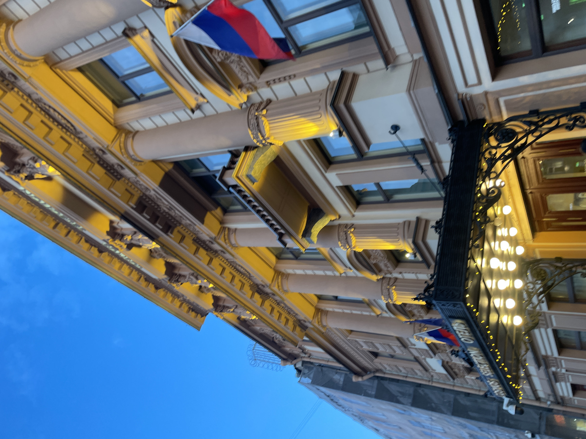 The image shows a low-angle view of a building facade in Saint Petersburg, Russia. The architecture is ornate, with classical elements such as columns, decorative moldings, and window frames. A Russian flag is visible, attached to the building's exterior. A black, wrought-iron sign adorned with illuminated light bulbs hangs from the building, displaying text in what appears to be a European language. The sky is visible in the background, transitioning from a dark blue to a lighter shade, indicating either dusk or dawn. The building's facade is lit with warm-toned lights.