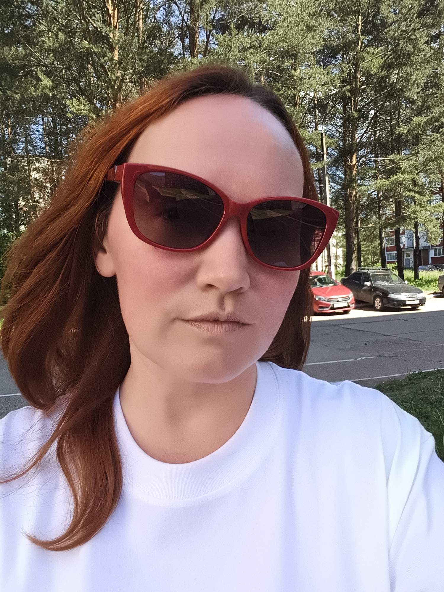 A medium shot captures a person with long, reddish-brown hair and red-framed sunglasses, wearing a white t-shirt. The person is outdoors, with a backdrop of tall, green coniferous trees. Behind the trees, a residential building with a red and white facade is visible, along with two parked cars, a red one and a dark gray one. The ground is paved, and there is green grass to the right. The scene appears to be in a residential area within the city of Chaikovsky, Russia, likely on a sunny day given the lighting.