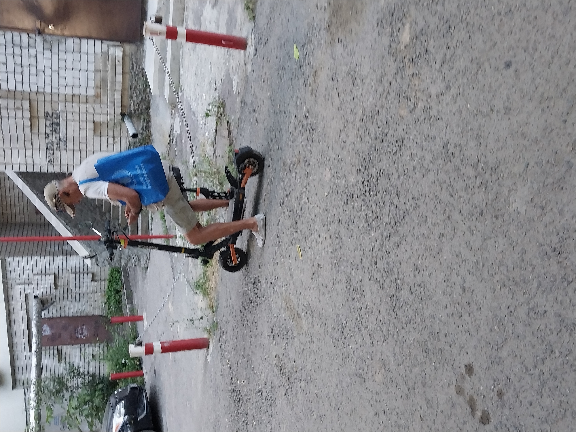 A man wearing a beige baseball cap, white t-shirt, and khaki shorts rides a black and orange electric scooter on a paved outdoor surface. He carries a large blue tote bag on his left shoulder. The scooter has a black frame and orange accents on its wheels and deck. He is wearing white slip-on shoes. The scooter appears to be stationary or moving slowly. In the background, a white brick building with a brown door is visible. Red and white bollards with chains are present in the foreground. The scene is set on a street in Dnipro, Ukraine.