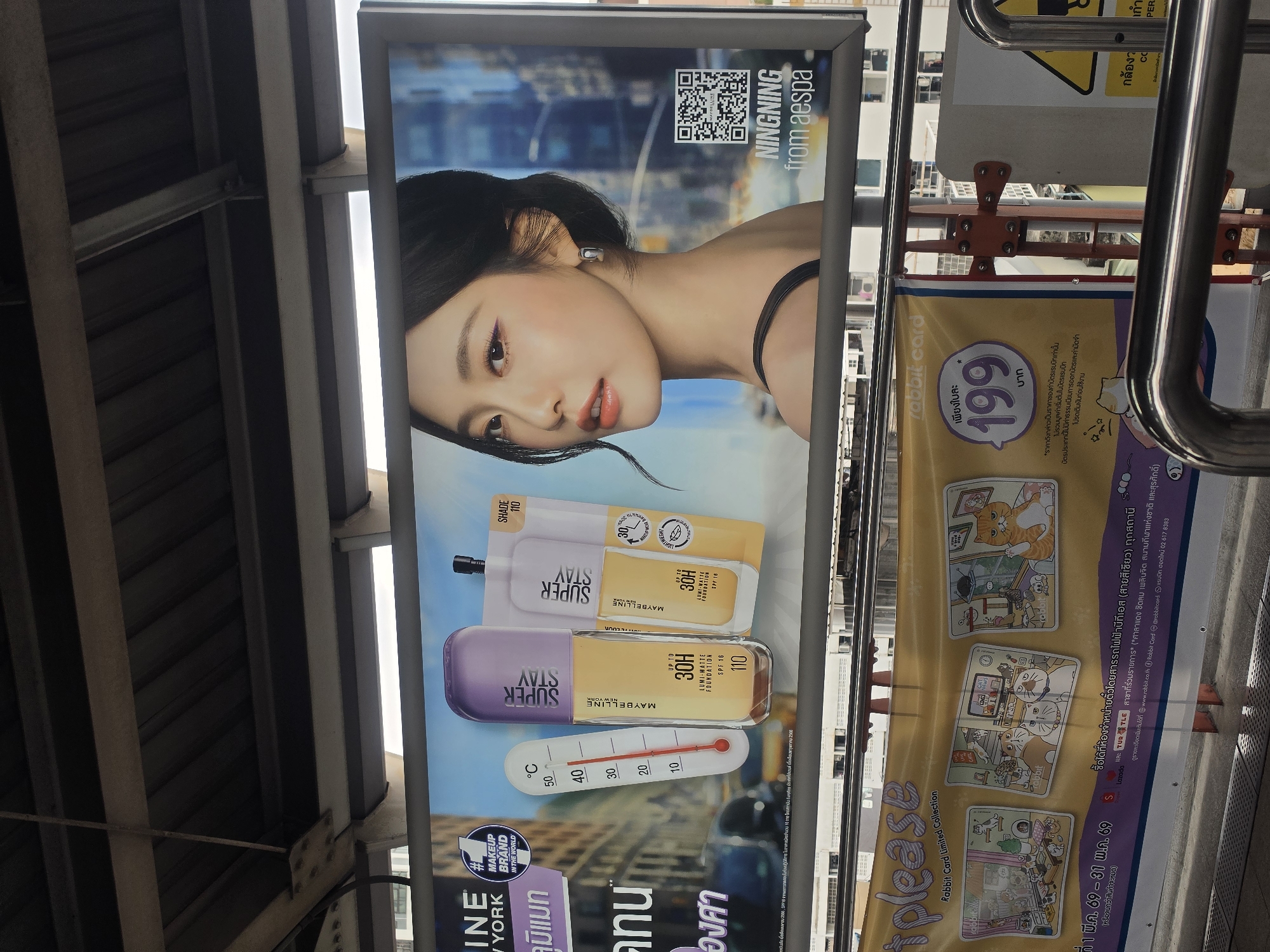 The image shows an advertisement for Maybelline Super Stay foundation, featuring Ningning from the South Korean girl group Aespa, displayed in a public transportation area in Bangkok, Thailand. The advertisement includes the products, a thermometer, and a QR code with the words "NINGNING from aespa". Next to the advertisement is a poster for Rabbit card, featuring cat illustrations, priced at 199 baht. The background shows metal structures typically found in elevated train stations.