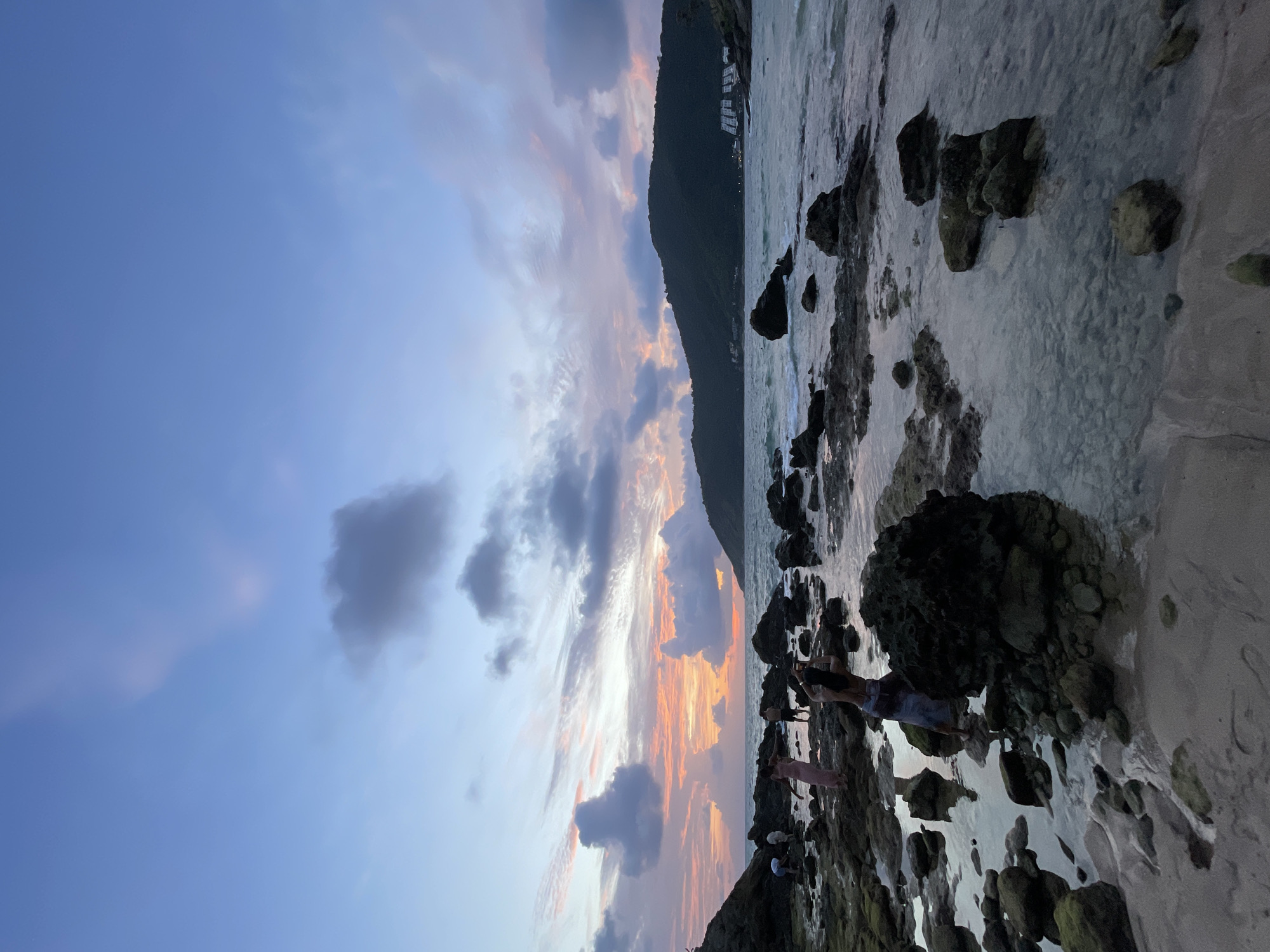 The image depicts a coastal scene in Rawai, Thailand, during what appears to be sunset. The sky is filled with a mix of soft blue, gray, and vibrant orange and pink hues, indicating the setting sun. Wispy clouds are scattered across the sky. In the background, a lush, dark green mountainside slopes down towards the sea. A few buildings are visible nestled on the mountain. The foreground is a rocky shoreline with clear, shallow water lapping over smooth stones and sand. Some larger, moss-covered rocks are present on the beach and in the water. Several people are visible on the shore, some interacting with the water and rocks, while others appear to be observing the sunset. One person is taking a photo with a phone. The overall mood is serene and peaceful, characteristic of a beach at dusk. There is no visible text in the image.