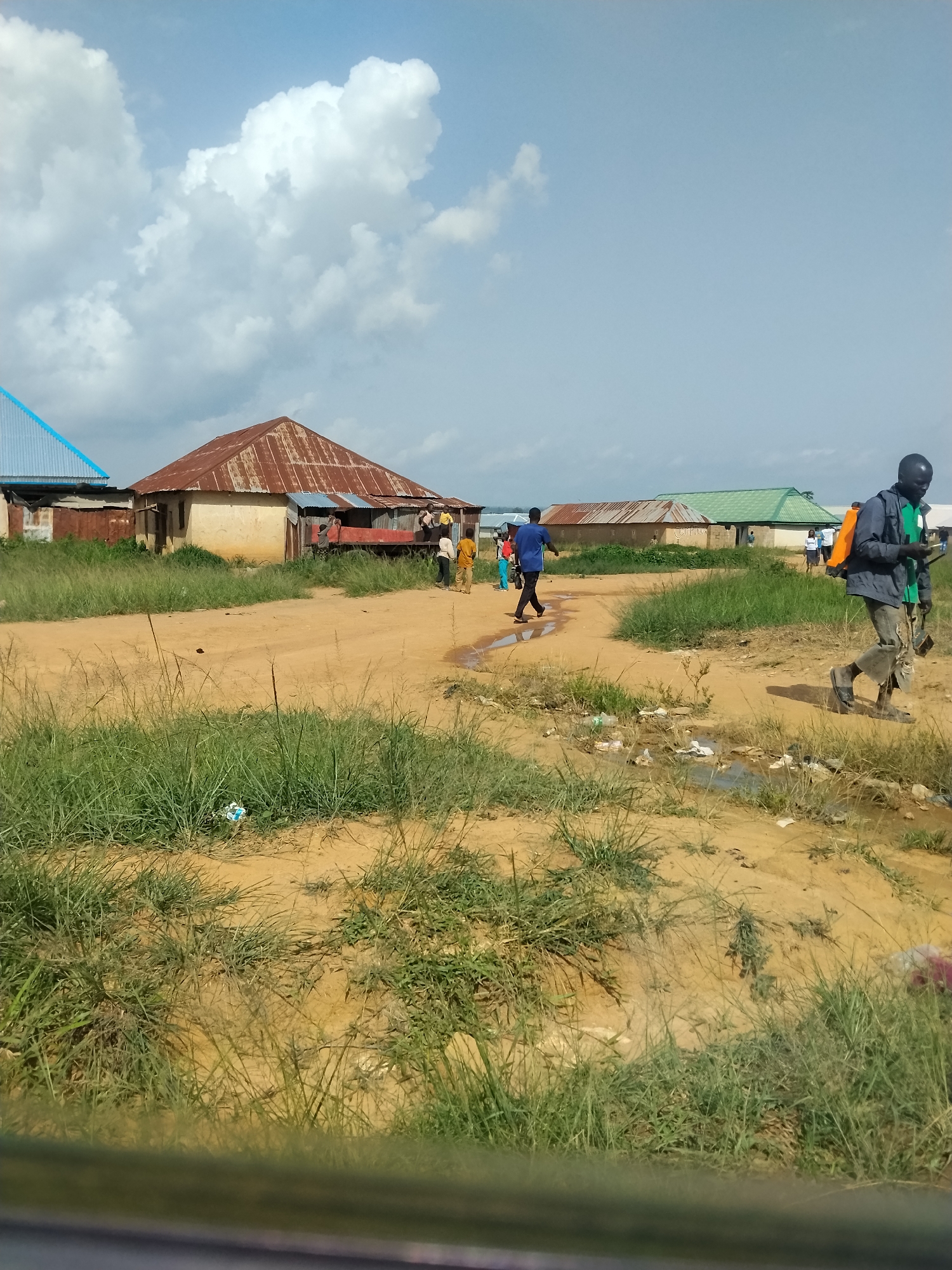 A group of people are walking on a dirt road in Durimi, Nigeria. The road is lined with grass and some scattered trash. In the background, there are several buildings with rusted metal roofs. The sky is blue with some white clouds. One person in the foreground is carrying a backpack and appears to be walking away from the camera. Another person in the distance is wearing a blue shirt and dark pants and is walking towards the camera.