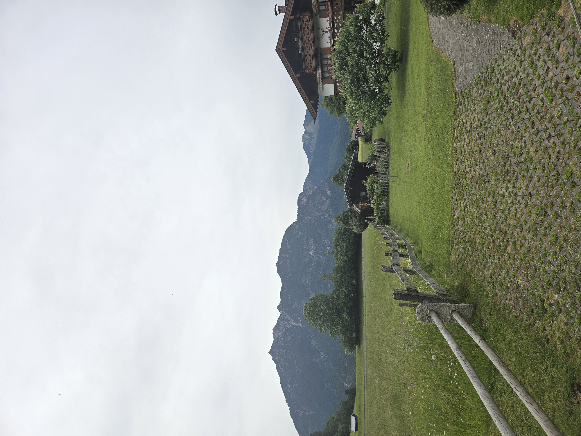 The image displays a tranquil mountainous landscape in what appears to be Garmisch-Partenkirchen, Germany, under an overcast sky. In the mid-ground on the right, a multi-story traditional Alpine-style house stands prominently. It features a dark pitched roof, white walls, and several wooden balconies adorned with intricate carvings and flowers in window boxes. Below it, a smaller building with a dark roof is nestled amongst trees.
Dominating the background are large, dark mountains with rugged peaks, partially obscured by the light grey, cloudy sky. The foreground and mid-ground consist of expansive, lush green lawns and fields. A rustic wooden fence, made of horizontal rails and vertical posts, runs horizontally across the mid-ground, extending towards the left. Closer to the viewer on the right side, a paved area is visible, featuring an irregular stone path that transitions into a section of grass pavers, where green grass grows through the grid-like concrete blocks. Several trees with dense green foliage are scattered throughout the scene, particularly around the buildings and at the base of the mountains.
The time of day appears to be daytime, though the overcast conditions prevent direct sunlight. No people are visible, and no specific activity is taking place, lending a peaceful and still atmosphere to the scene. There is no discernible text in the image.
