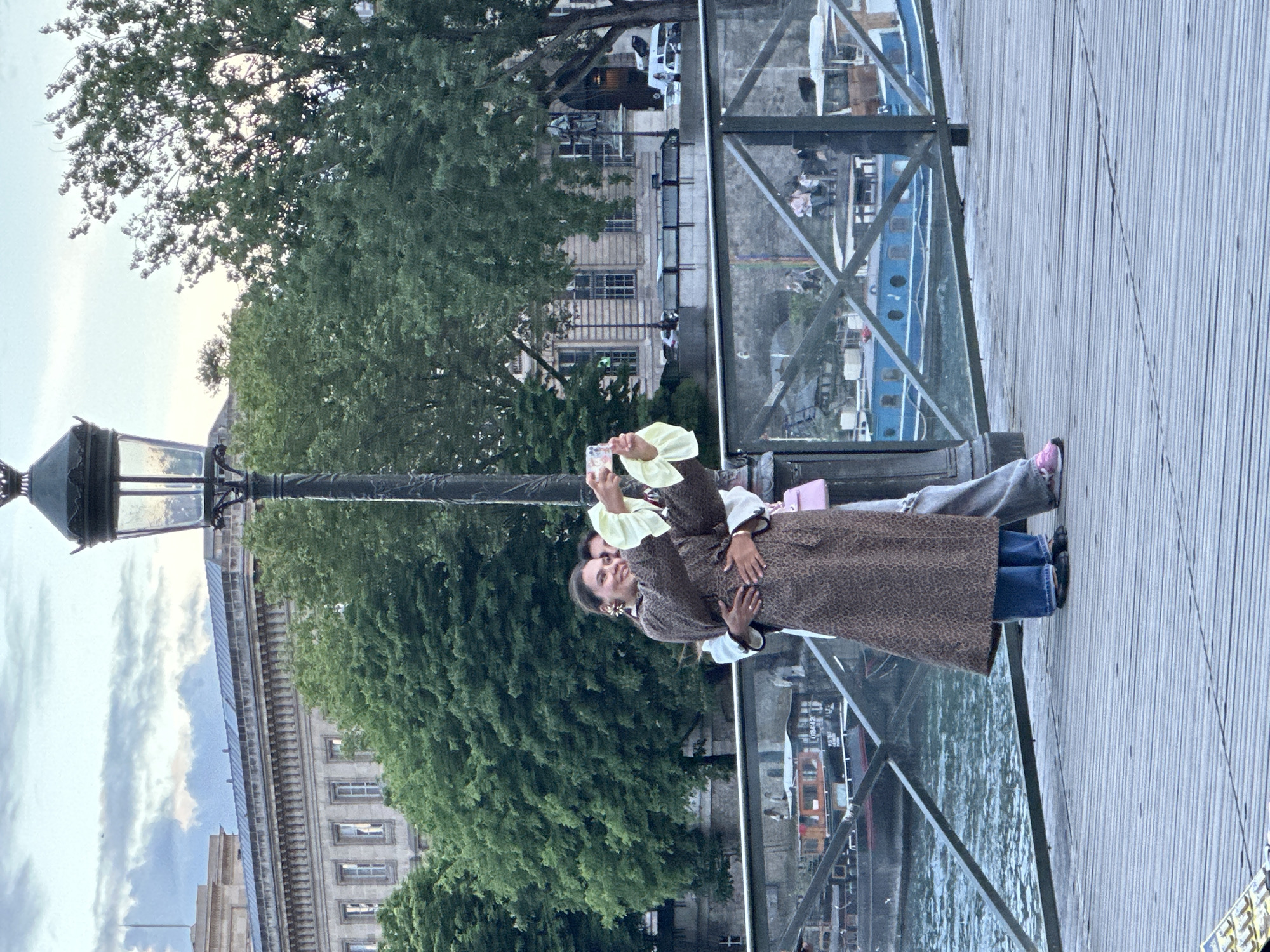 Two people stand closely together on a wooden walkway, a woman in a leopard-print coat holding up a phone to take a picture. A river is beside them with a boat in the background. The Pont des Arts bridge is visible, indicating this scene is in Paris, France. Trees and buildings line the river. A lamppost stands nearby.