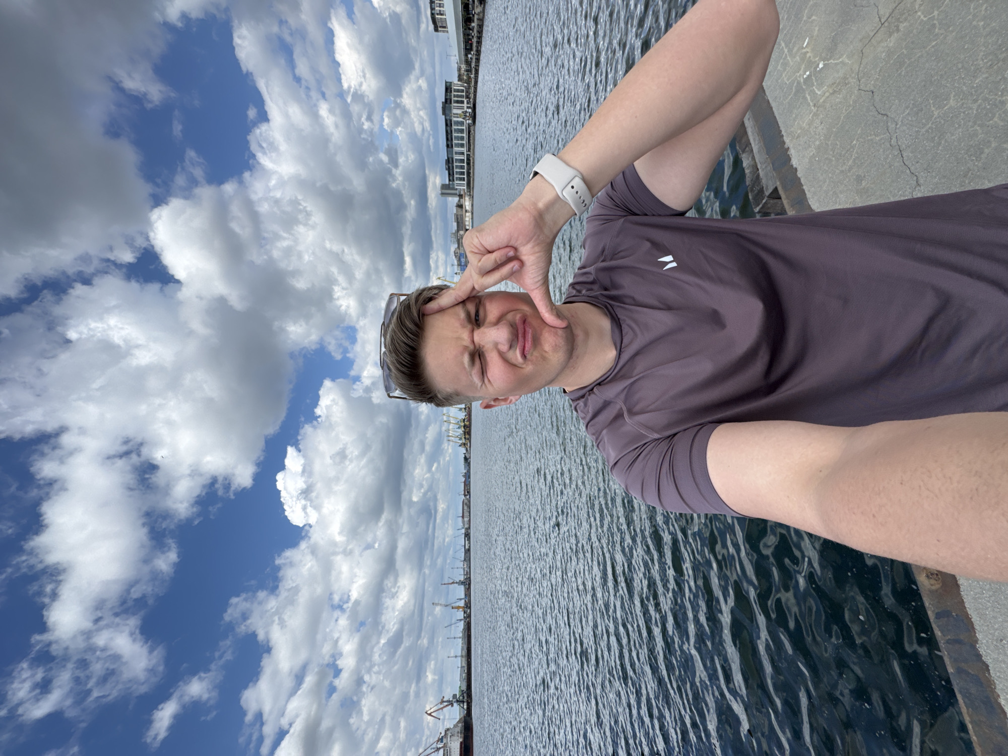 A man takes a selfie on the quay in Burgas, Bulgaria. The man, fair-skinned with short blonde hair and wearing a purple t-shirt and a white smartwatch, makes a square frame with his fingers around his face. Sunglasses are tucked into the collar of his shirt. Behind him is the sea, with various ships and cranes visible in the distance. The sky is filled with fluffy white clouds. A grey concrete barrier is located to the right. Buildings are visible in the distance to the left.