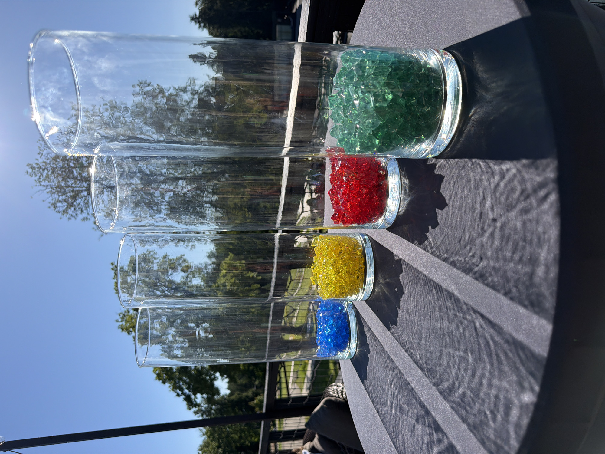 Four tall, clear glass vases are arranged in a line on a dark gray table outdoors. Each vase is filled with colored glass gems at the bottom. From left to right, the vases contain blue, yellow, red, and green glass gems. The vases are reflecting the sunlight and the surrounding trees, which are visible in the background against a bright blue sky. A black metal railing and a hint of outdoor furniture are also visible in the background. The image appears to be taken on a sunny day. The city of Peresvet, Russia is not identifiable from the image.