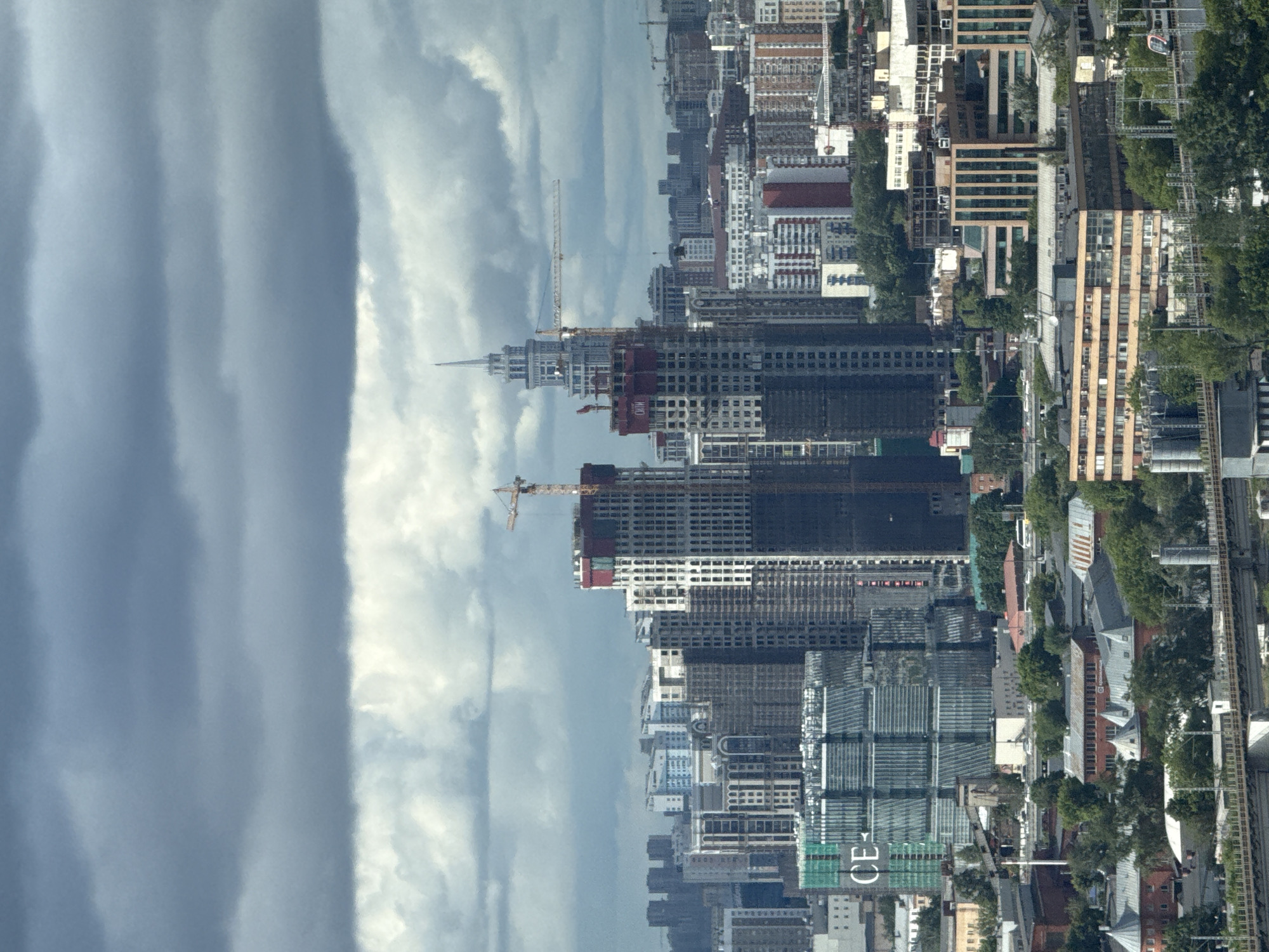 The image depicts a cityscape, likely in Khimki, Russia, given the prompt's specification. The perspective is an elevated view, possibly from a high-rise building. The scene showcases a dense urban environment with numerous buildings, including several under construction. Construction cranes are visible near the unfinished structures. Completed buildings display diverse architectural styles, ranging from modern glass-clad towers to more traditional designs. A railway track runs along the right side of the image, parallel to a tree-lined street. The sky is overcast with dramatic cloud formations. The general impression is of an area undergoing rapid development and modernization.