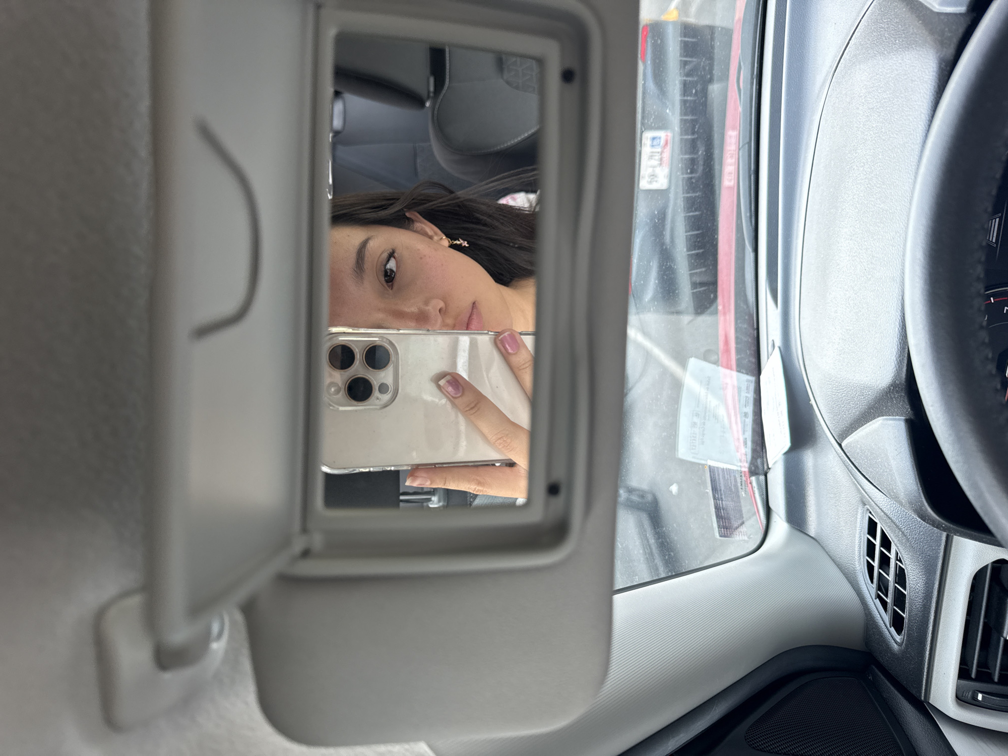 A woman is reflected in the mirror of a car's sun visor. The mirror is reflecting her holding a smartphone, likely taking a selfie. Outside the car window, one can see the edge of another vehicle, with a license plate visible. The license plate appears to be from Puebla, Mexico, with partial characters visible: "59-L20." The interior of the car is visible, including the steering wheel and dashboard elements.