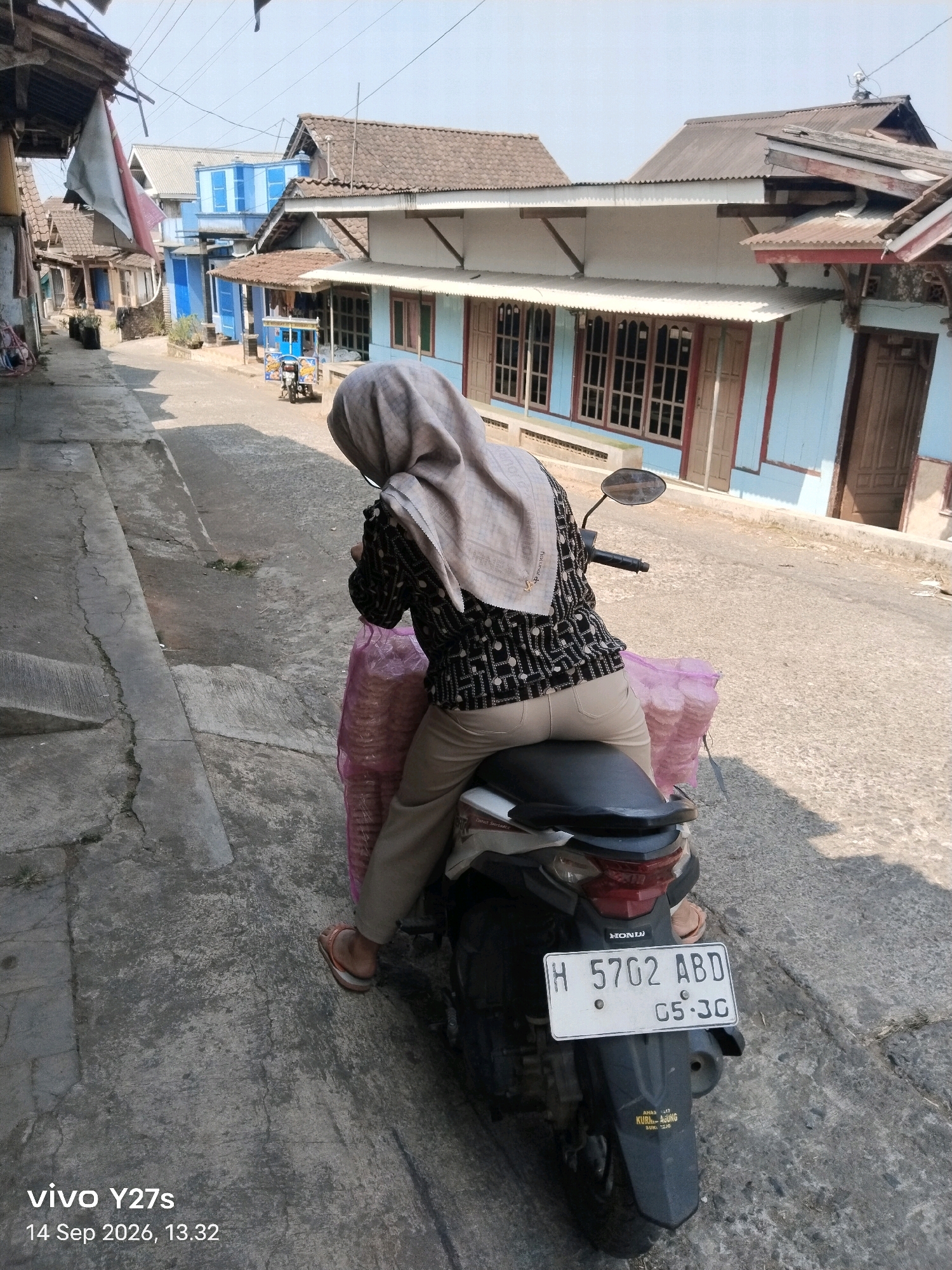 A woman wearing a light-colored hijab, a black and white patterned shirt, and tan pants is sitting on a scooter in an outdoor, urban environment. She is facing away from the camera, and the scooter is parked on a paved street. A large, pink plastic bag filled with what appears to be snacks or food items is secured to the back of the scooter. The scooter has a license plate that reads "H 5702 ABD 05-36". In the background, there are several buildings with light blue and white paint, some with brown tiled roofs, and a narrow street lined with these structures. The scene appears to be in a village or town in Tamansari, Indonesia, given the architecture and the motorcycle's license plate format, which suggests an Indonesian registration. The weather is sunny, casting shadows on the ground. The timestamp on the image indicates it was taken on September 14, 2026, at 1:32 PM.