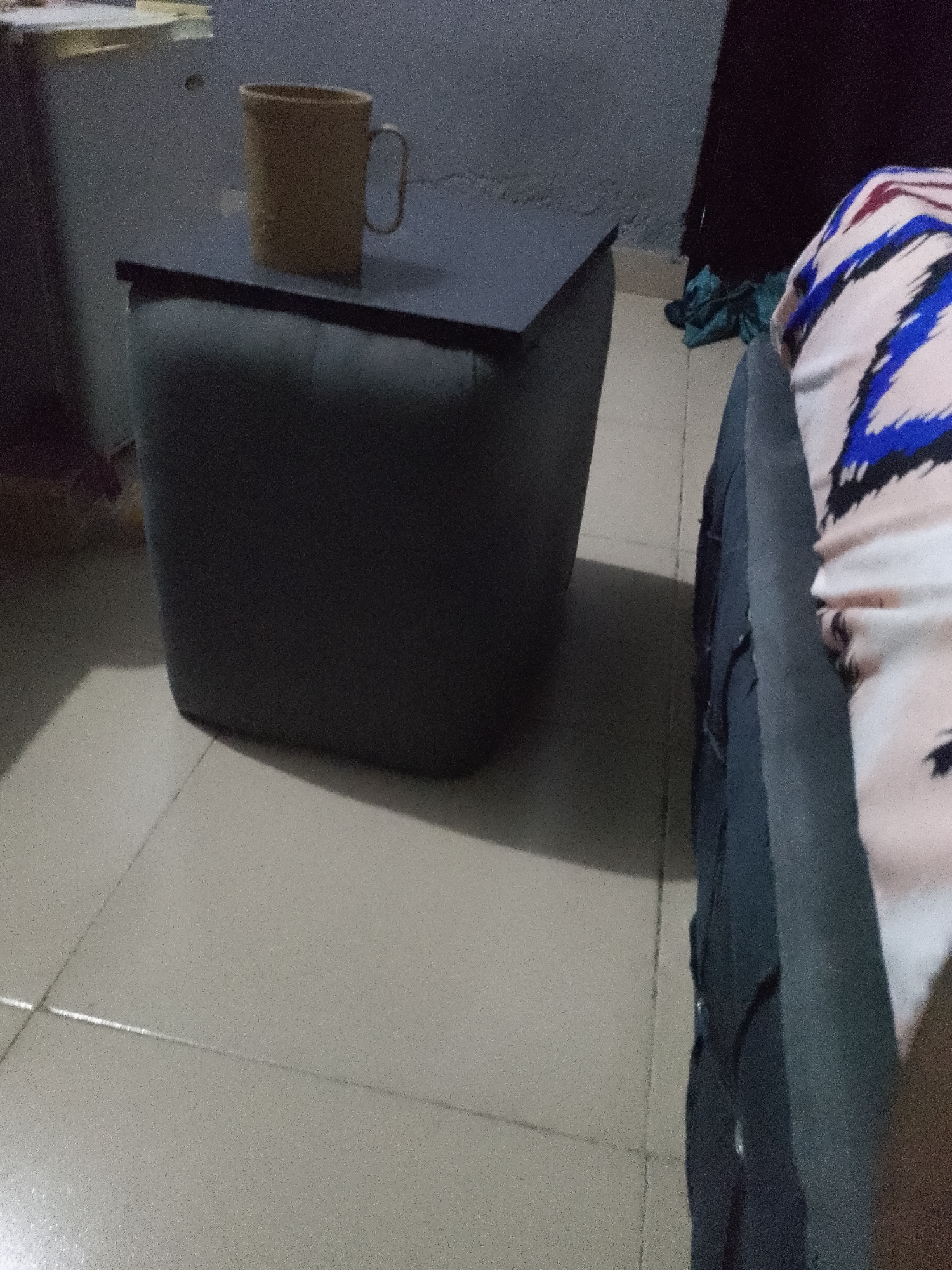 A beige mug with a handle sits on a dark gray cube-shaped table in a dimly lit room. The table has a dark gray top and a padded, dark gray body. The floor is tiled with light beige square tiles. To the right of the table, a portion of a person wearing a white, pink, blue, and black patterned garment is visible. The background is dark and out of focus. The scene appears to be indoors, possibly a bedroom or living area.