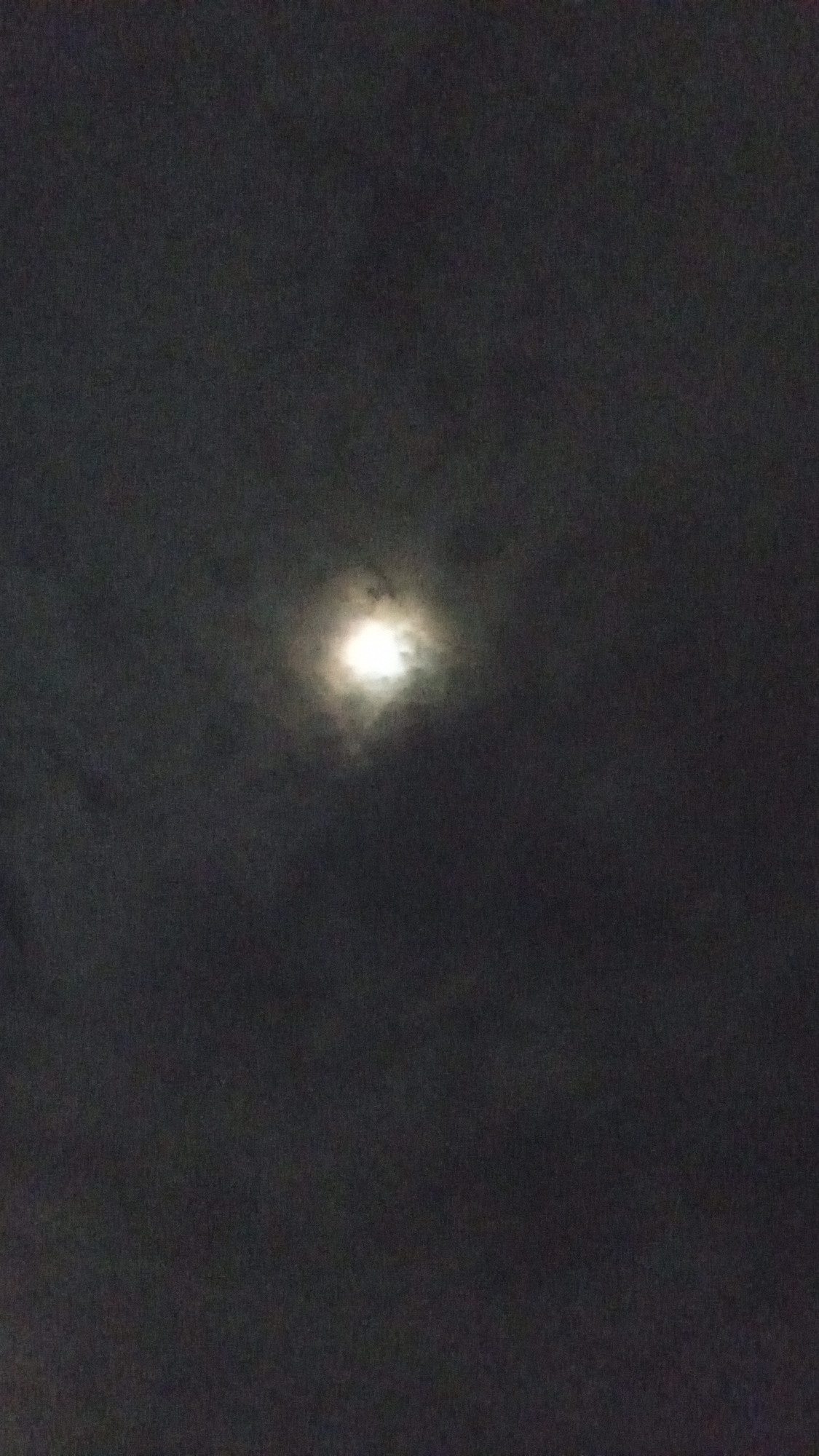 The image shows a night sky with a bright moon partially obscured by clouds. The environment is dark, with varying shades of gray representing the clouds and the overall night sky. The moon appears as a luminous, circular shape surrounded by a slightly hazy halo caused by the diffusion of light through the clouds. No people, subjects, objects, or actions are visible, making it difficult to assess more context about the scene, except its general outdoor environment. The precise location within Buketteukuh, Indonesia, cannot be determined without additional visual or contextual cues.